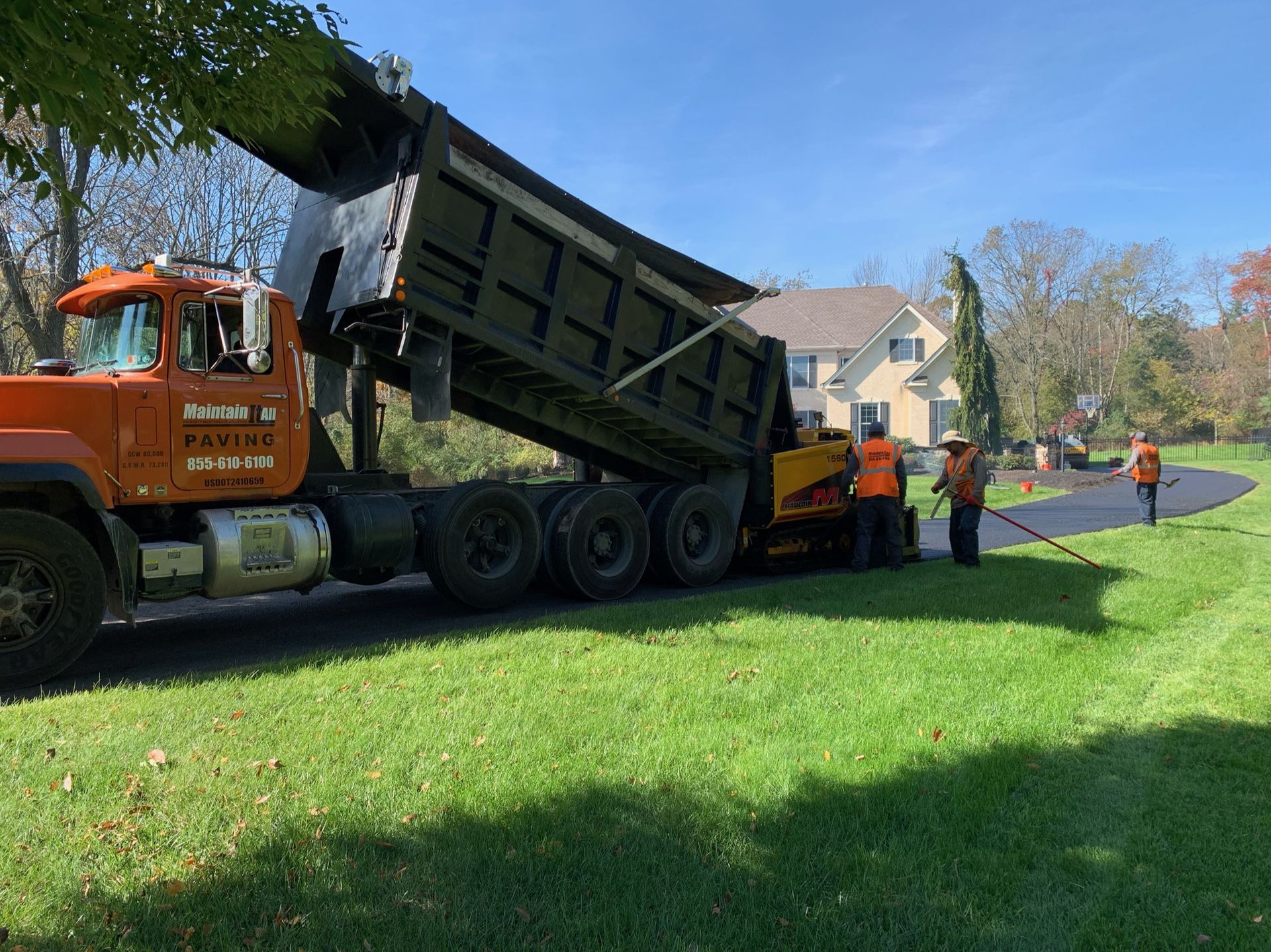 A dump truck with its bed raised pours asphalt into a paving machine as workers in orange safety vests operate the equipment.