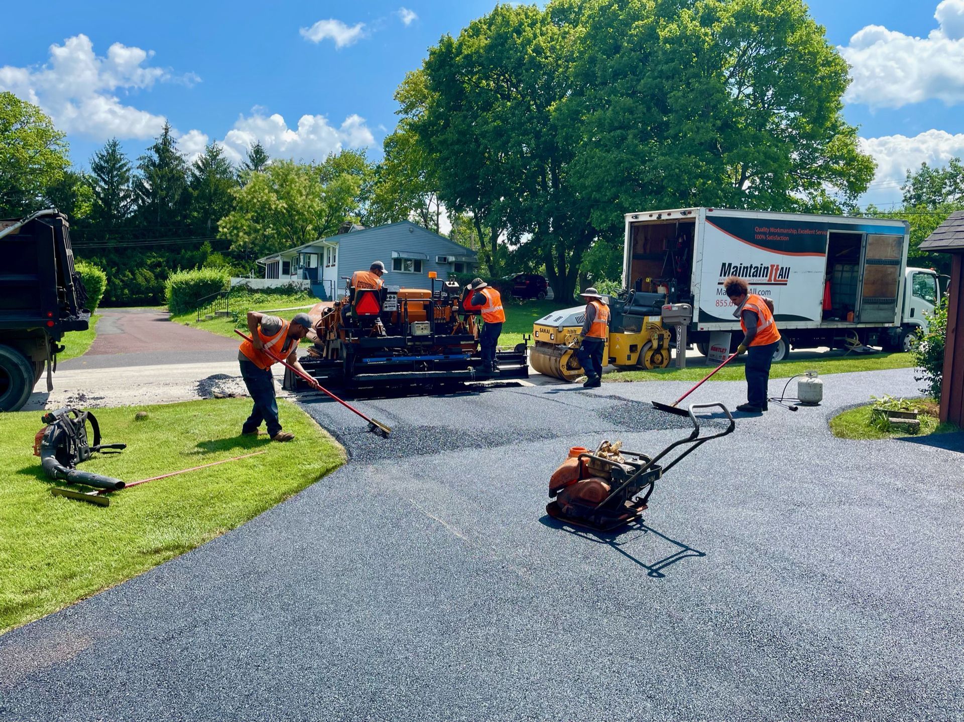 A construction crew in orange high-visibility vests paves a residential driveway with asphalt on a sunny day.