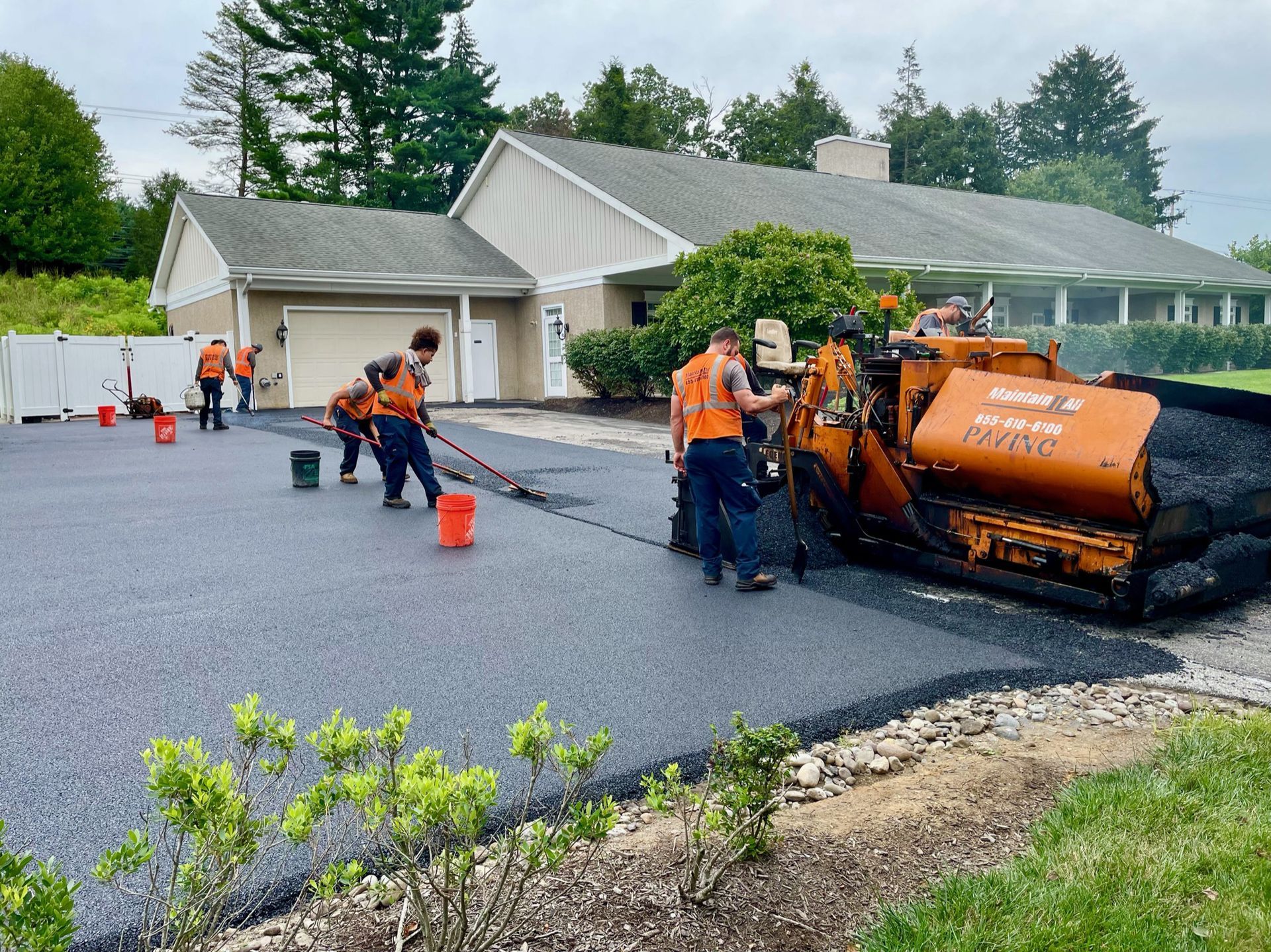 A crew in orange safety vests paves a residential driveway using heavy machinery and tools on a sunny day.