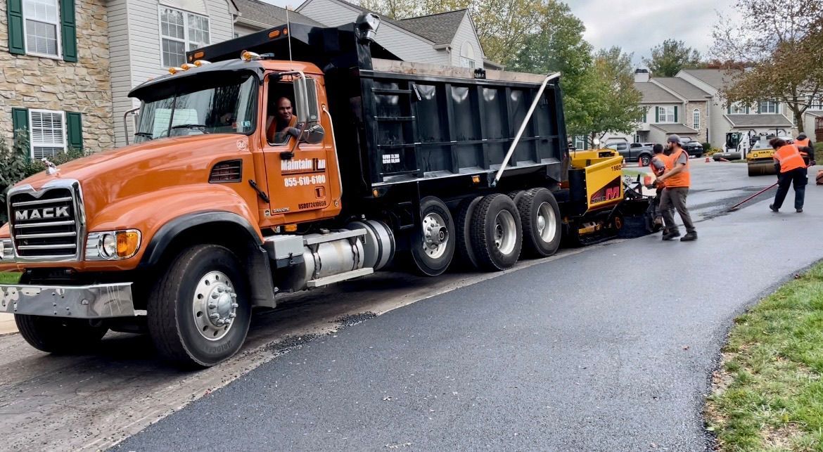 An orange dump truck and yellow paver work on a suburban street as crew members lay fresh black asphalt.