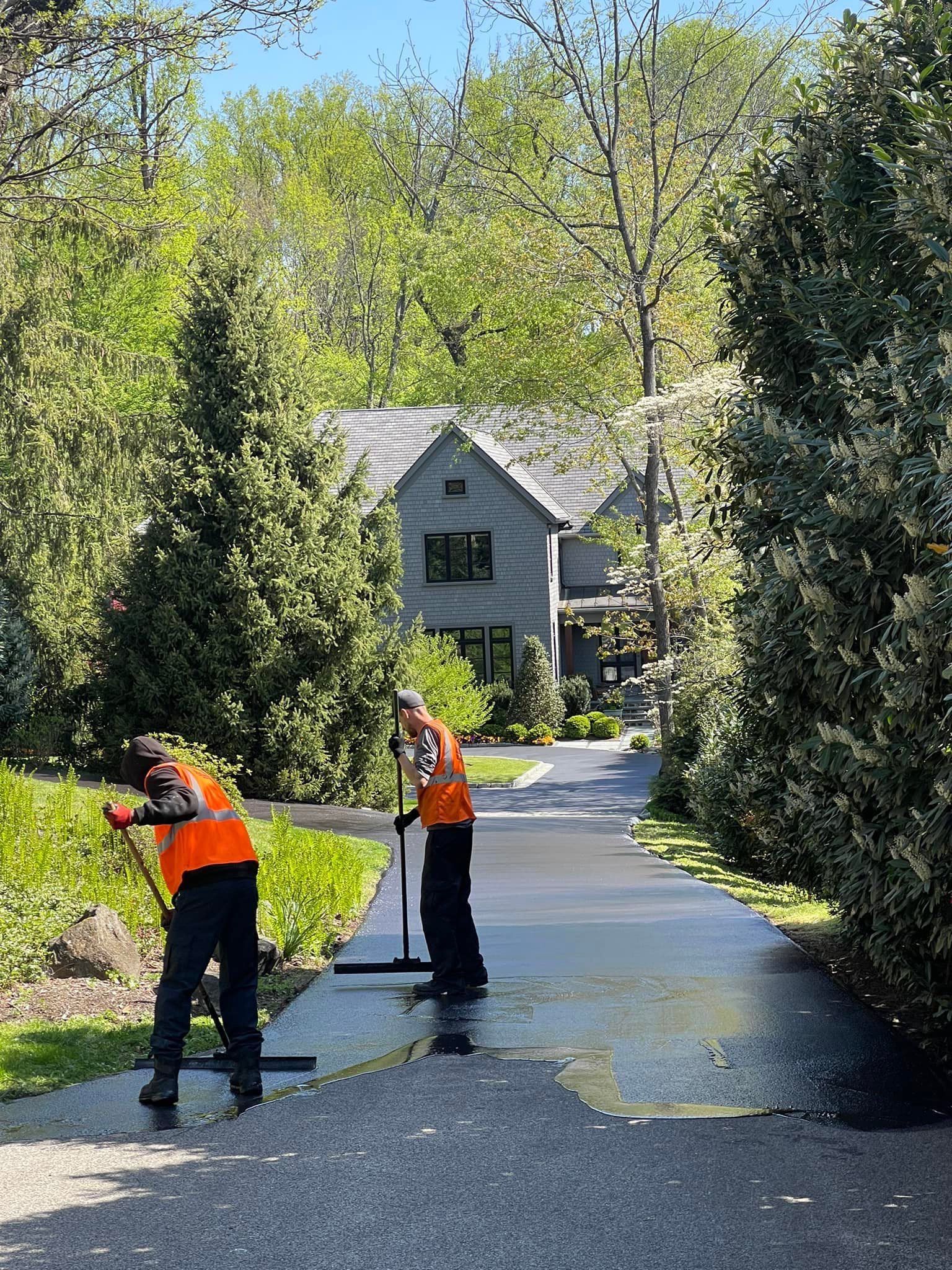 Two workers in bright orange vests use squeegees to apply dark sealcoat to a residential asphalt driveway.