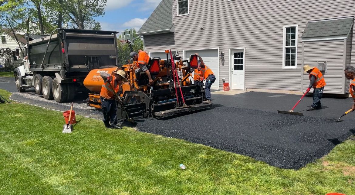 Construction workers in safety vests use machinery to pave a driveway with fresh black asphalt next to a house.