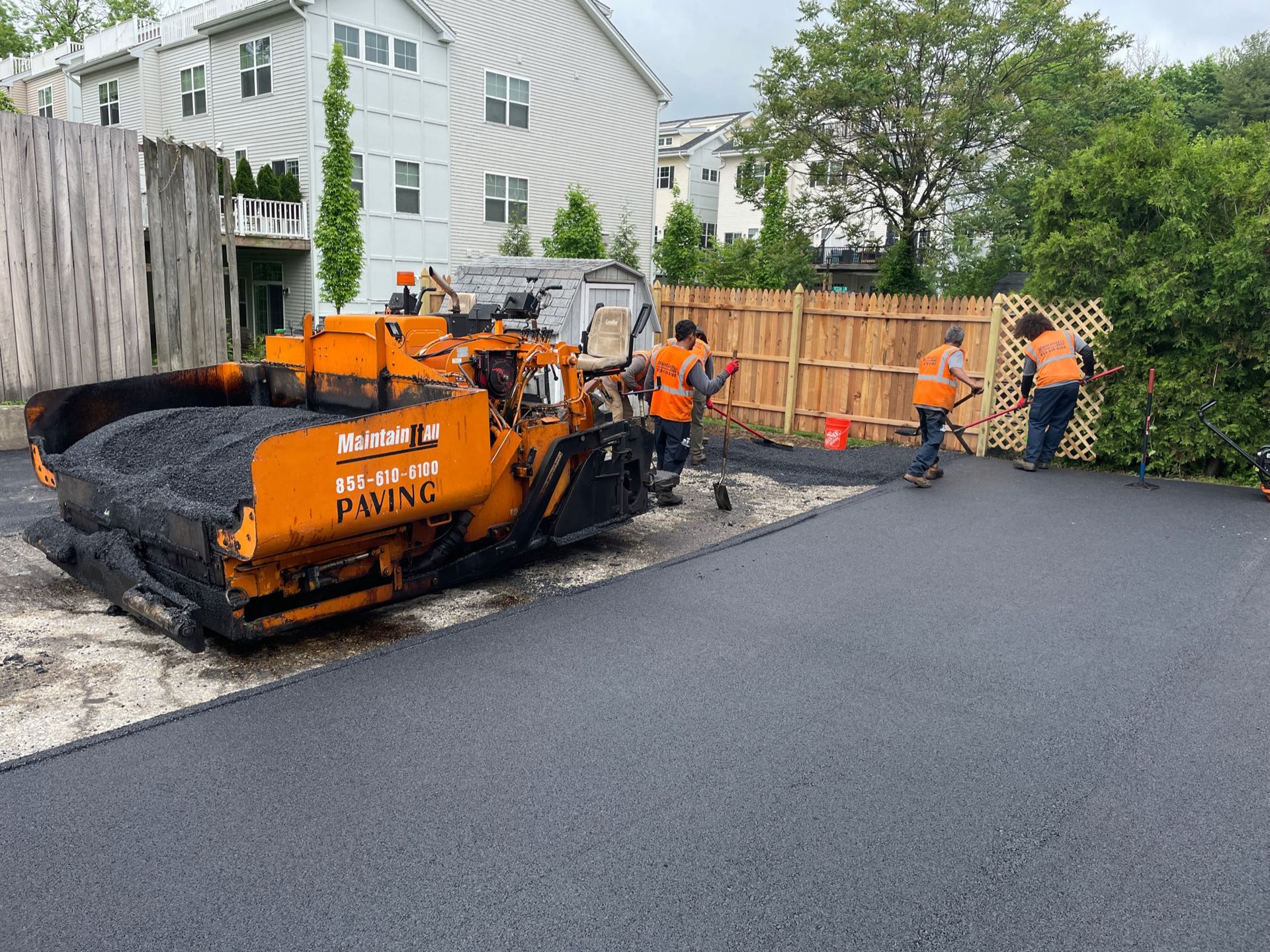 Workers in orange safety vests operate an asphalt paving machine on a suburban driveway construction site.