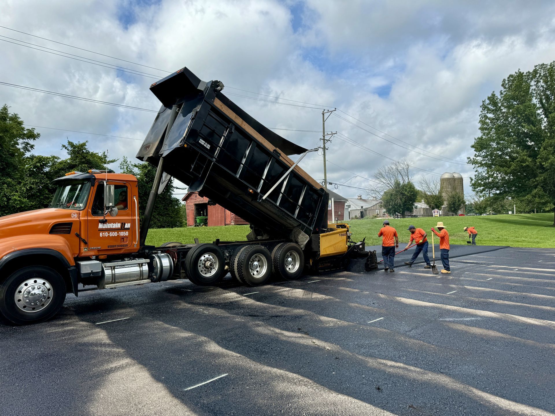 An orange dump truck pours asphalt onto a parking lot while a road crew spreads it out in a sunny outdoor setting.