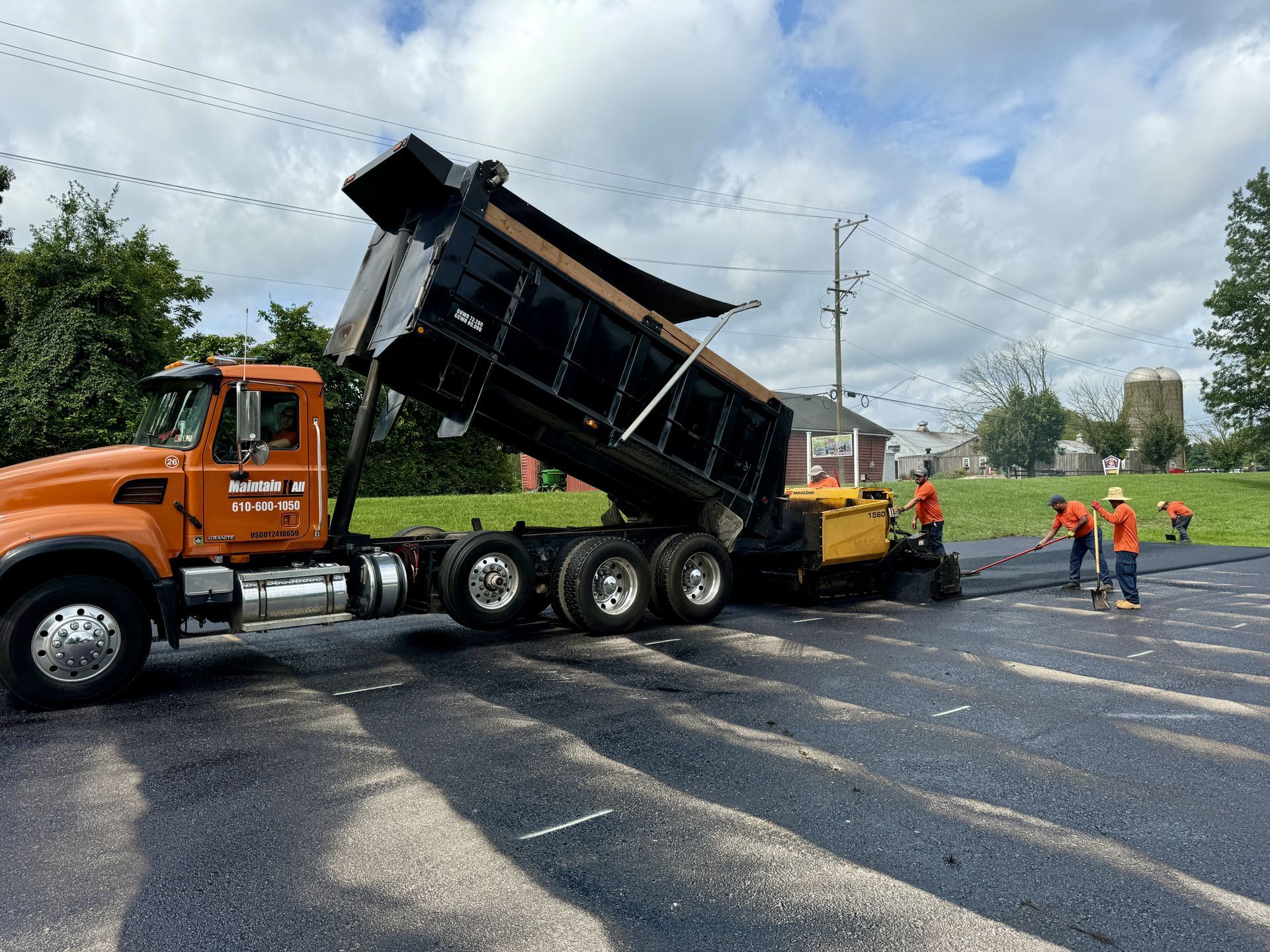 A dump truck pours asphalt into a paving machine on a sunny day as road workers stand nearby.