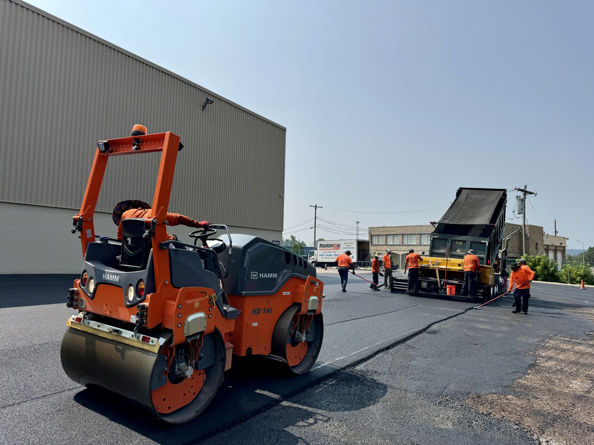 Construction workers paving a road with an orange asphalt roller and a yellow paving machine on a sunny day.