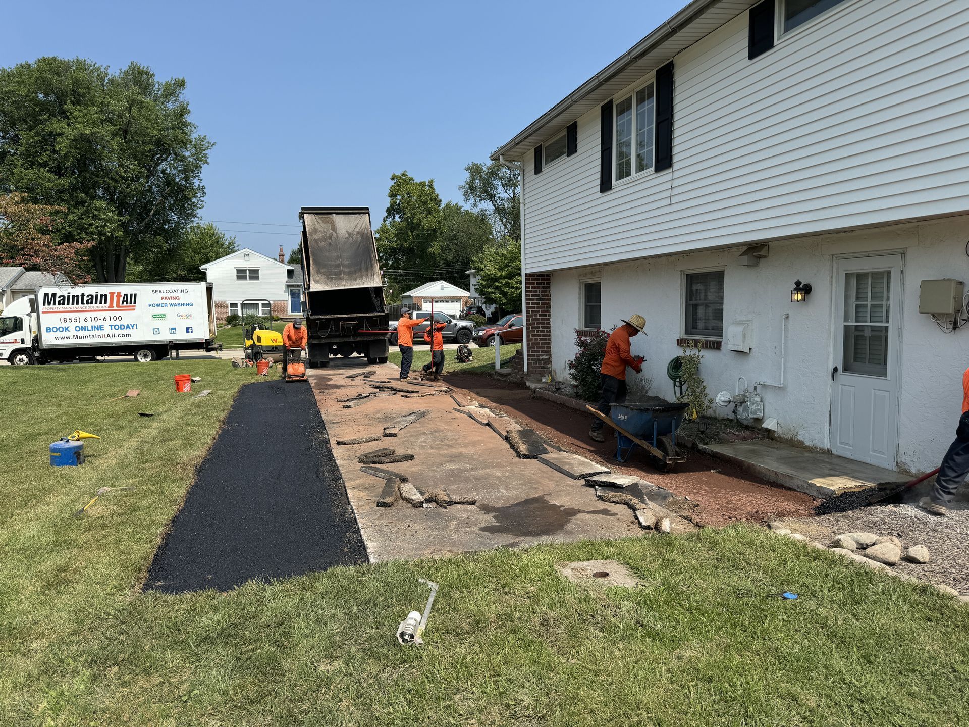 Workers pave a residential driveway with fresh black asphalt next to a white house on a sunny day.