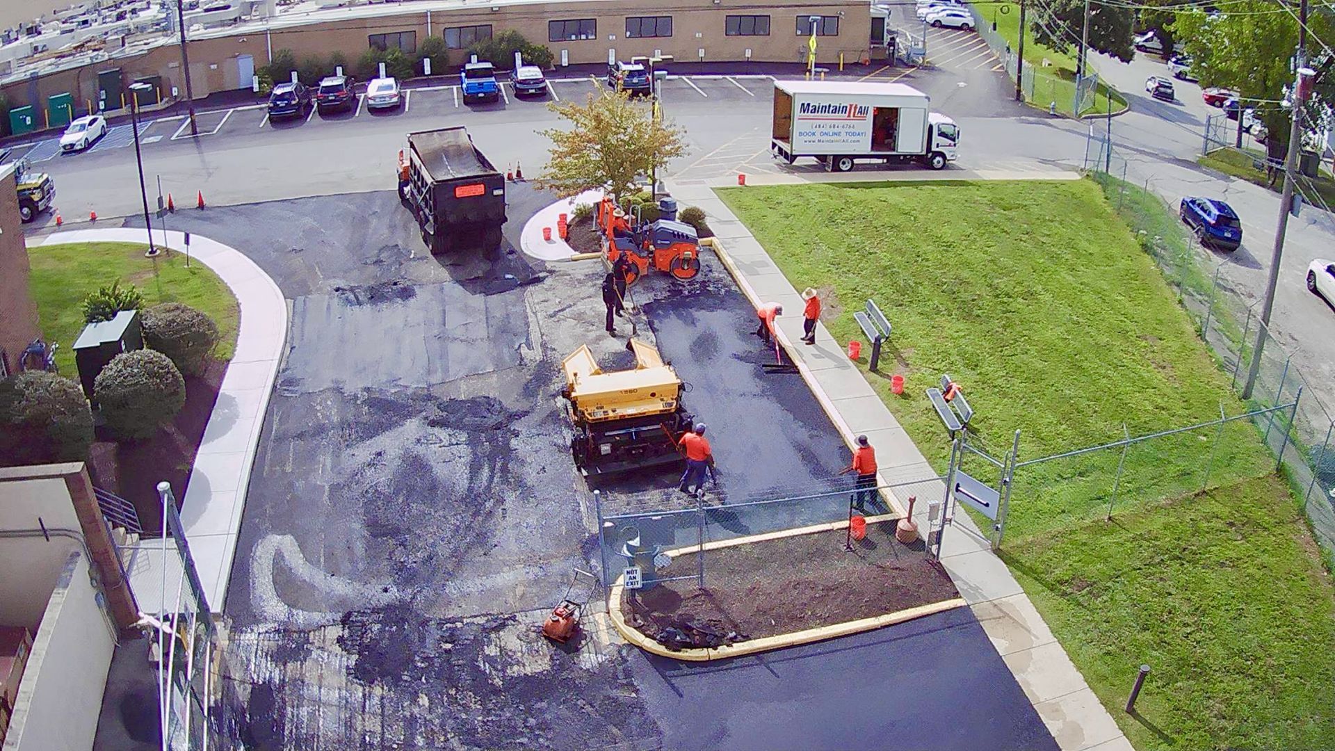 A high-angle view of a crew paving a parking lot with machinery and dump trucks near a grassy area and commercial building.