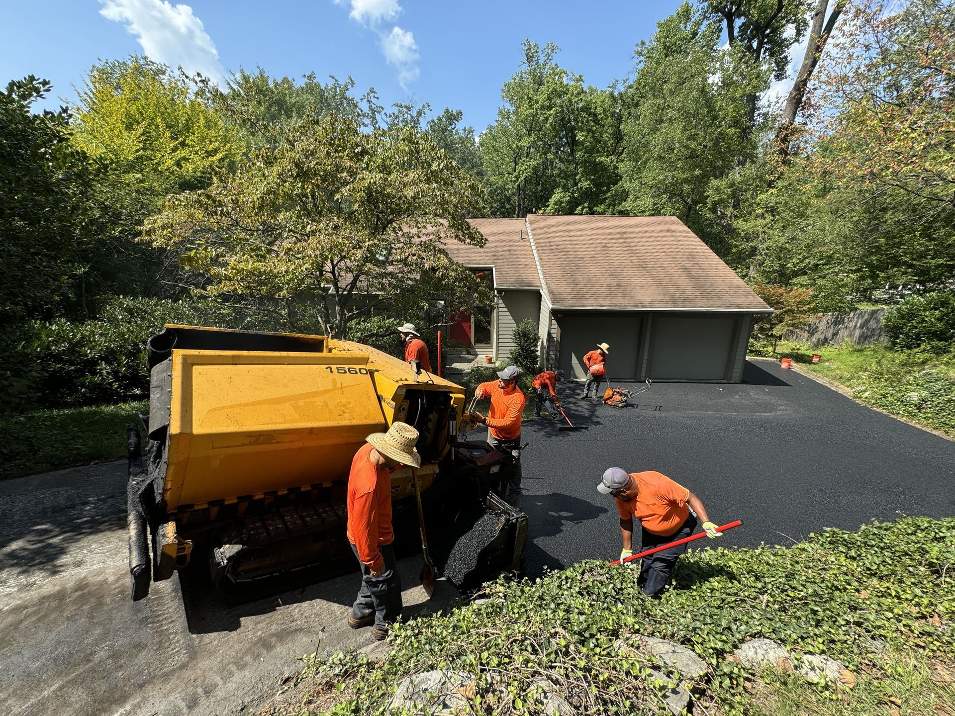 Construction workers in orange shirts operate a yellow paving machine to resurface a driveway in front of a house.