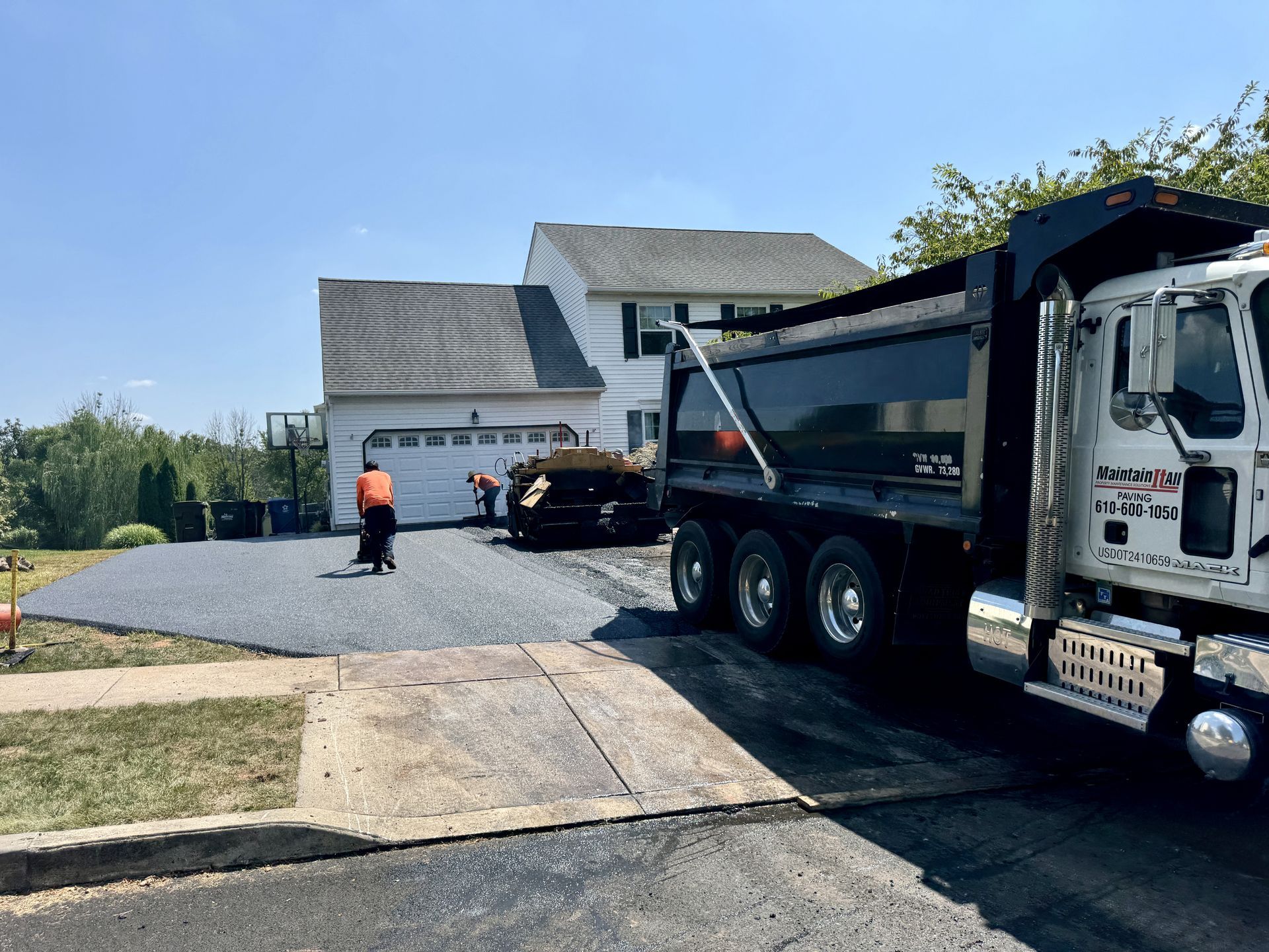 A dump truck parked on a residential driveway as construction workers pave the surface with gravel.