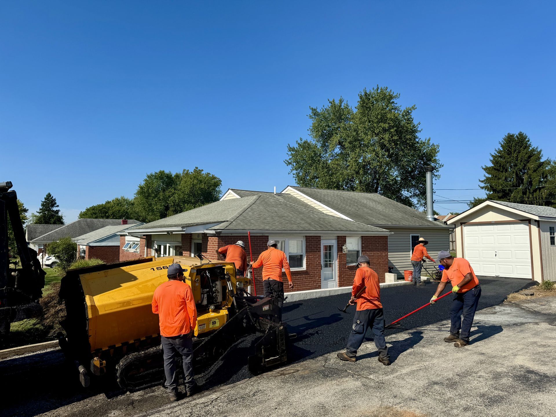 Workers in orange safety vests pave a driveway with a yellow machine in front of a house on a sunny day.