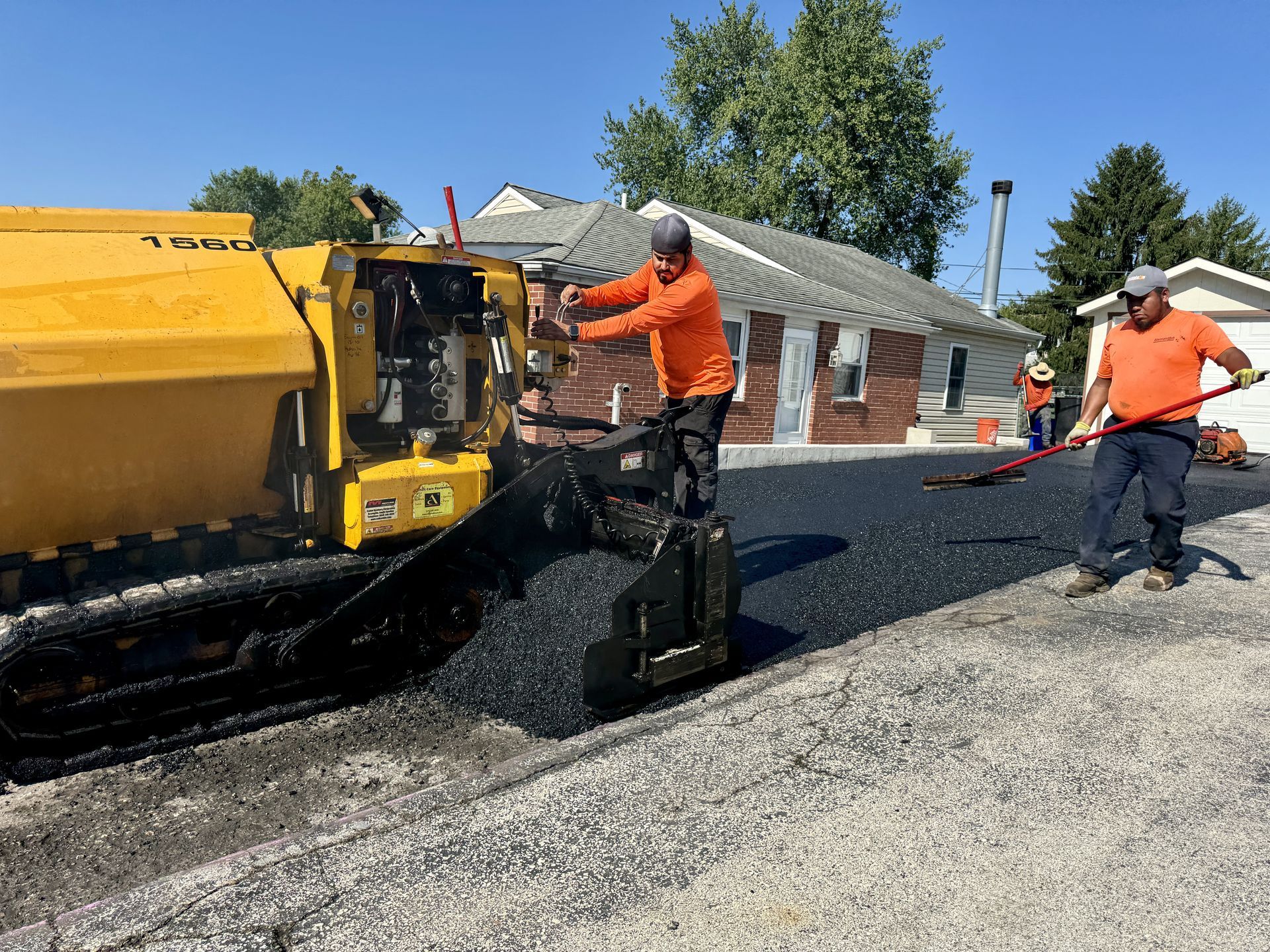 Two workers in orange shirts operate construction machinery to lay fresh asphalt on a driveway outside a house.