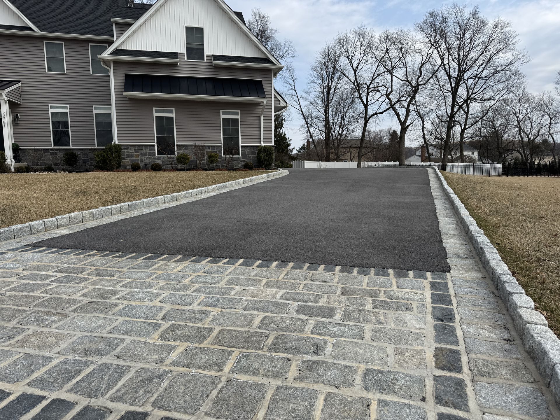 A gray house with a stone-bordered asphalt driveway extending from the foreground into the yard.
