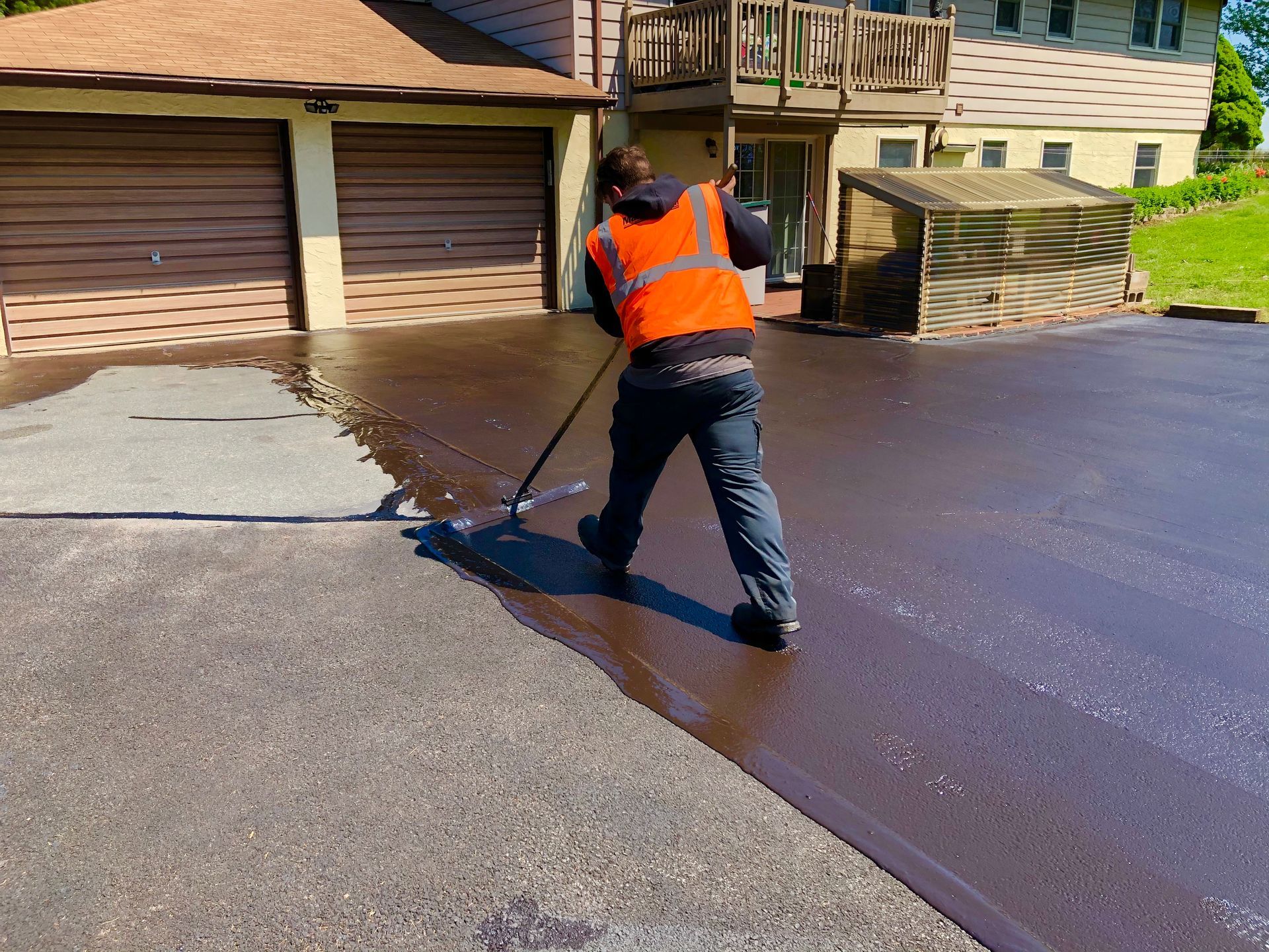 A person in a high-visibility vest uses a squeegee to apply black sealant to a residential driveway.
