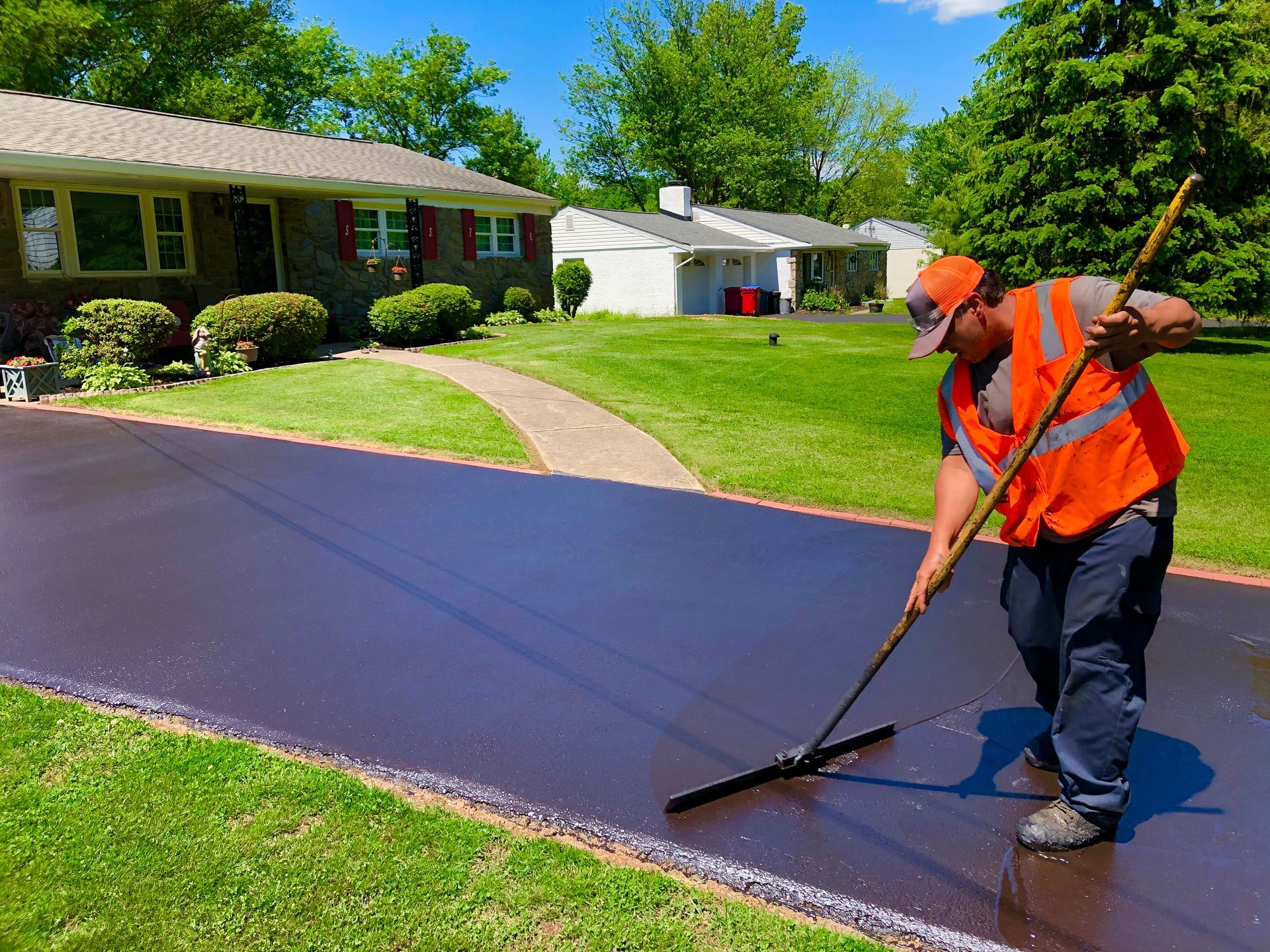 A person in an orange safety vest uses a squeegee to apply dark asphalt sealer to a residential driveway.