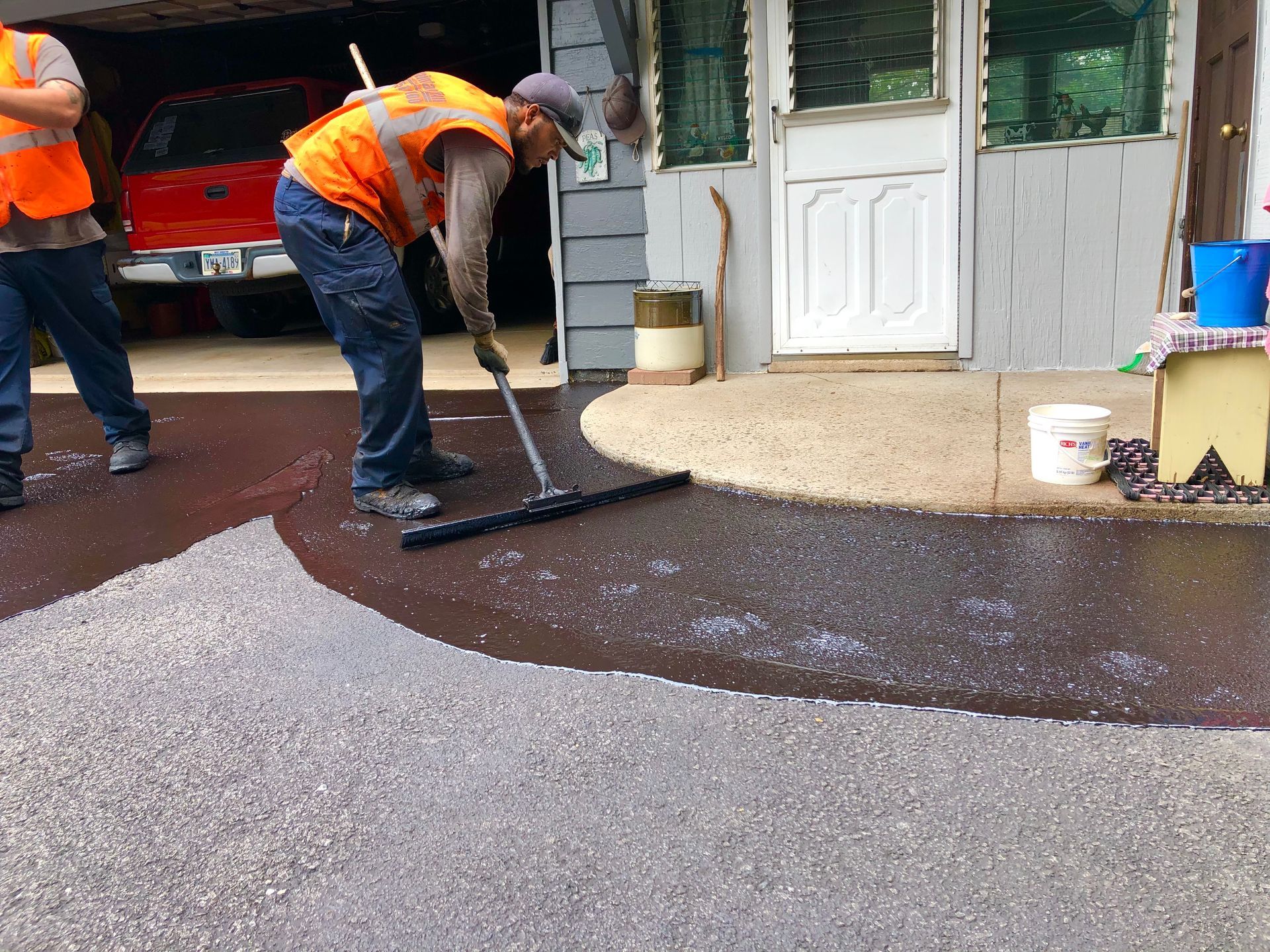 Two workers in safety vests apply black driveway sealant with a squeegee in front of a house.