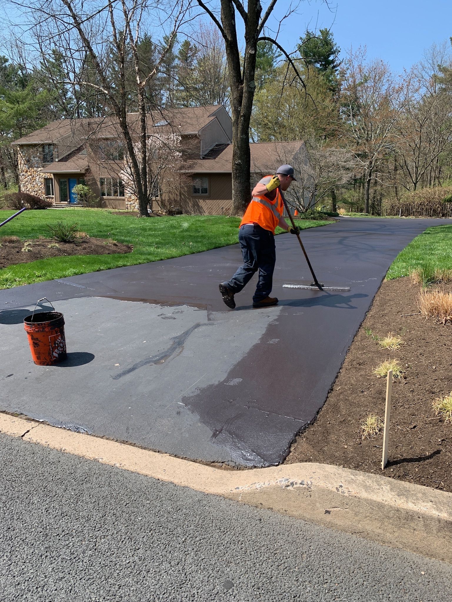 A worker in an orange vest uses a long-handled tool to apply sealant to a residential asphalt driveway.