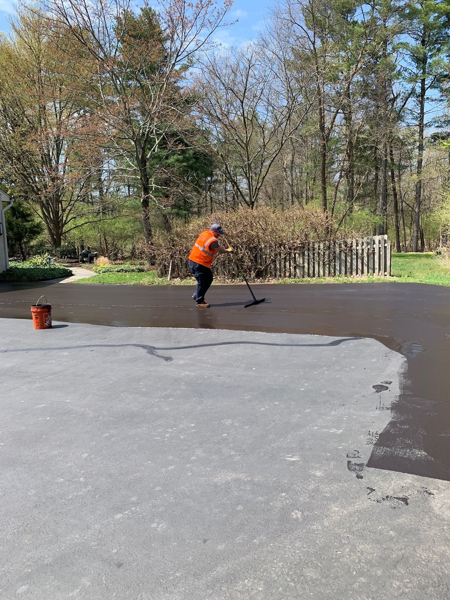 A person in an orange shirt uses a squeegee to apply black sealant to a gray asphalt driveway.