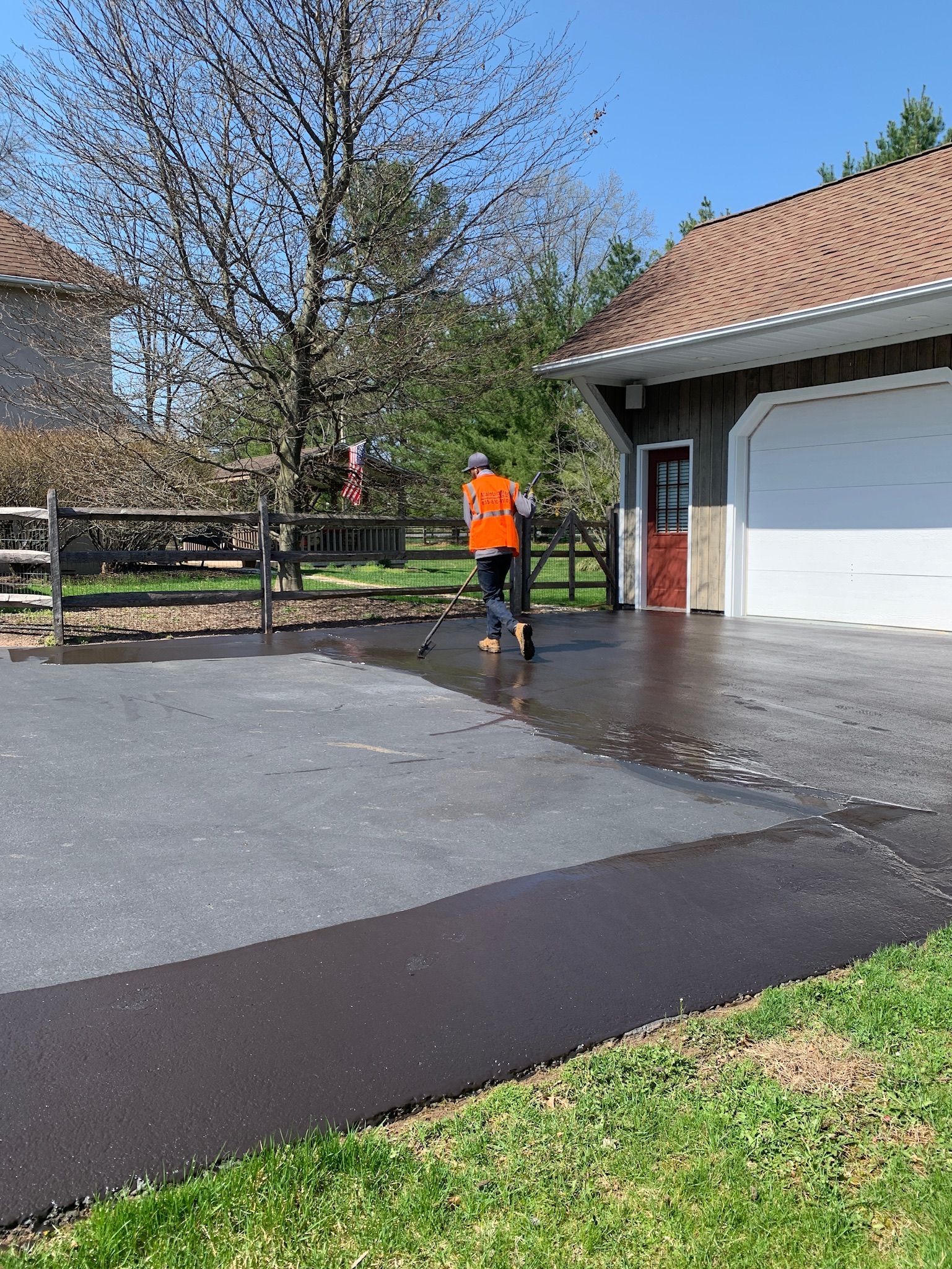 A person in a bright orange safety vest applies black sealant to a driveway in front of a garage and a tree-lined yard.