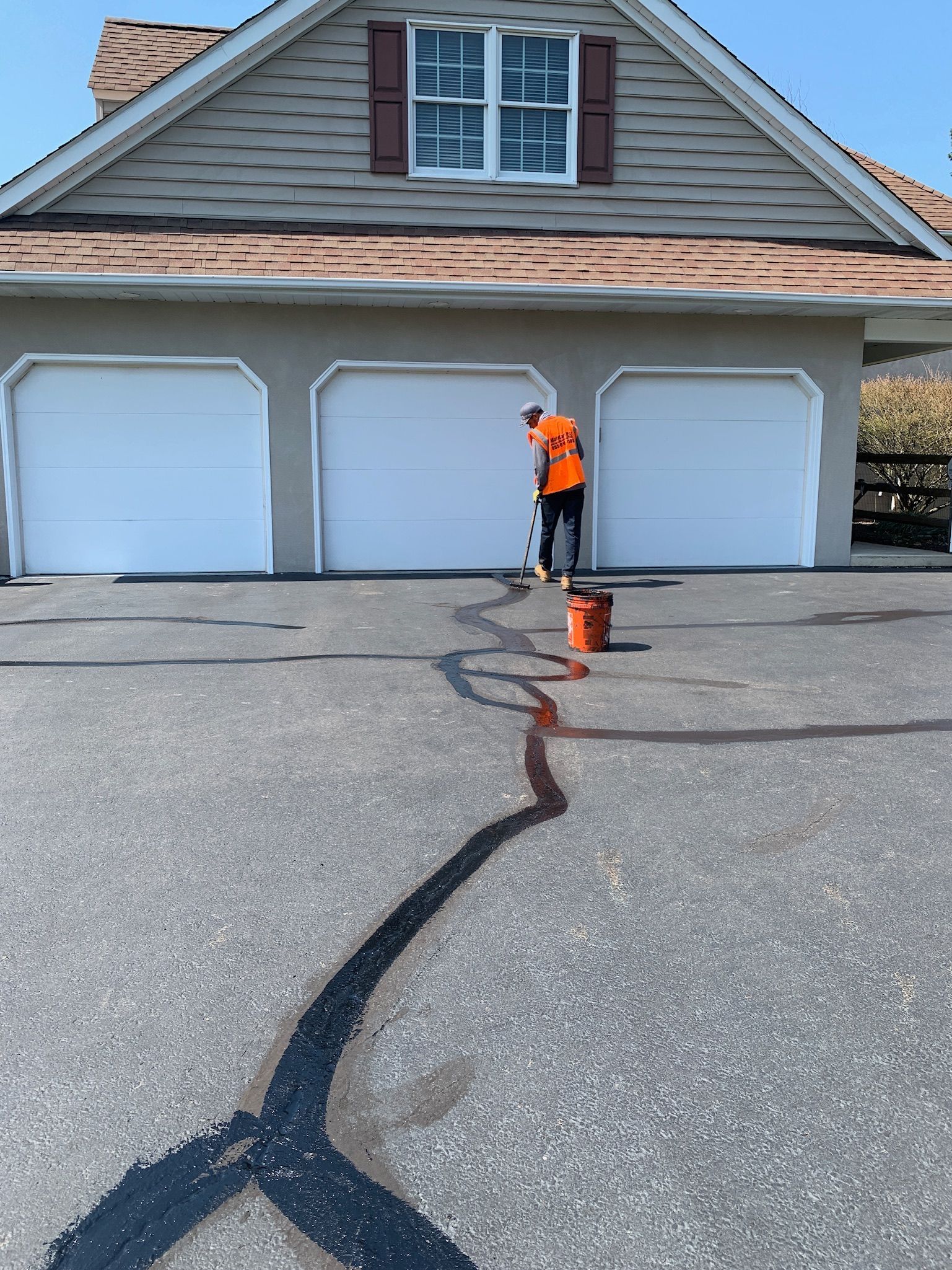 A person in a bright orange safety vest fills cracks in an asphalt driveway with black sealant in front of a house.
