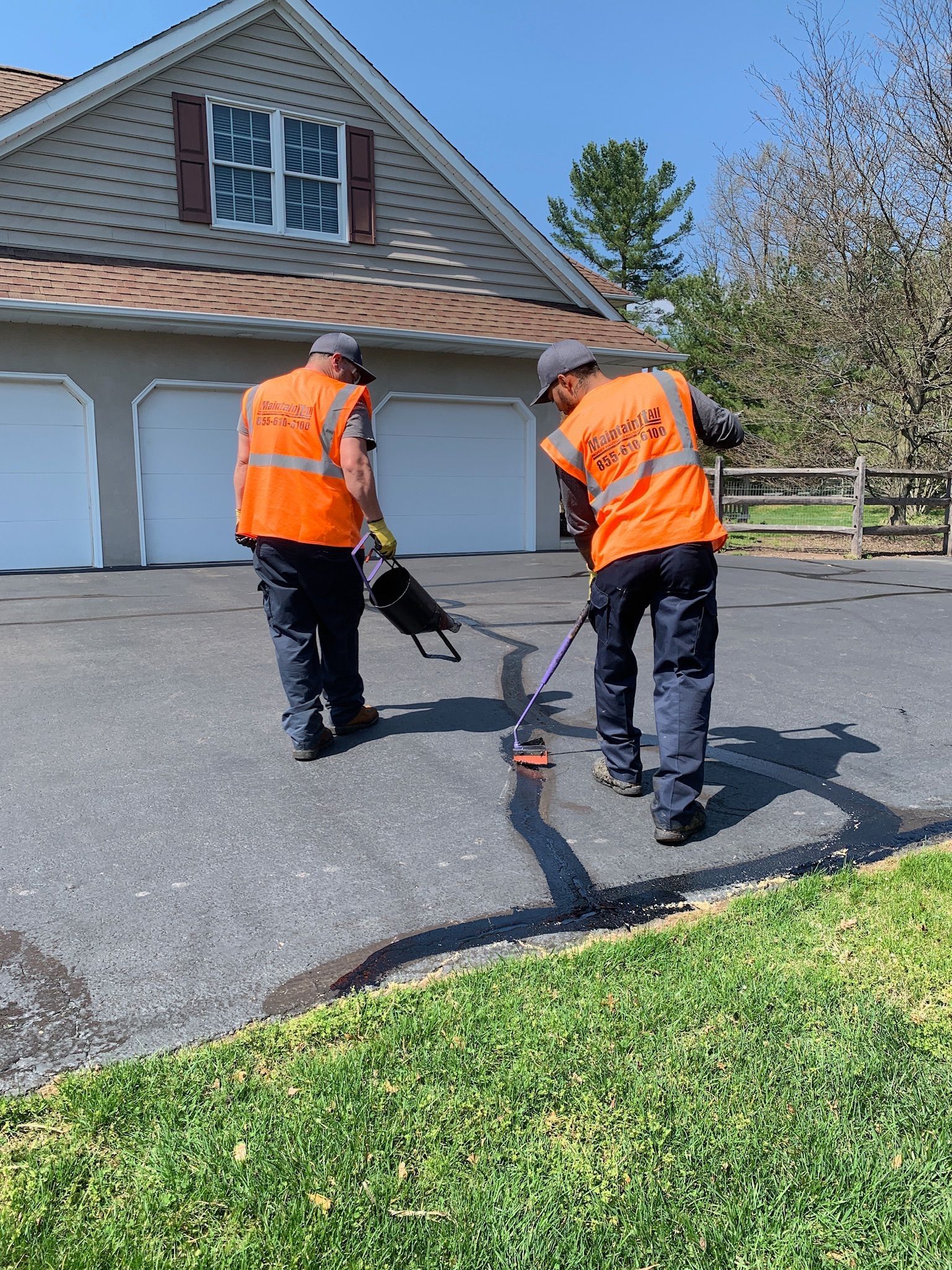 Two workers in orange safety vests repair cracks on an asphalt driveway in front of a residential garage.
