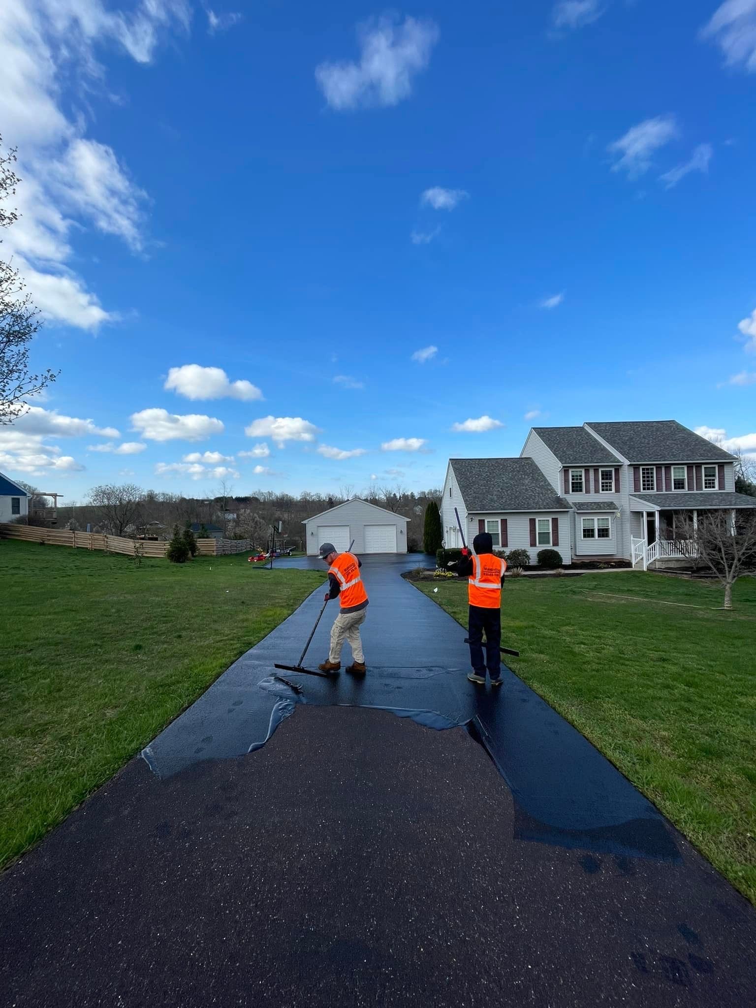 Two workers in orange safety vests use long brushes to apply black sealant to a residential driveway.