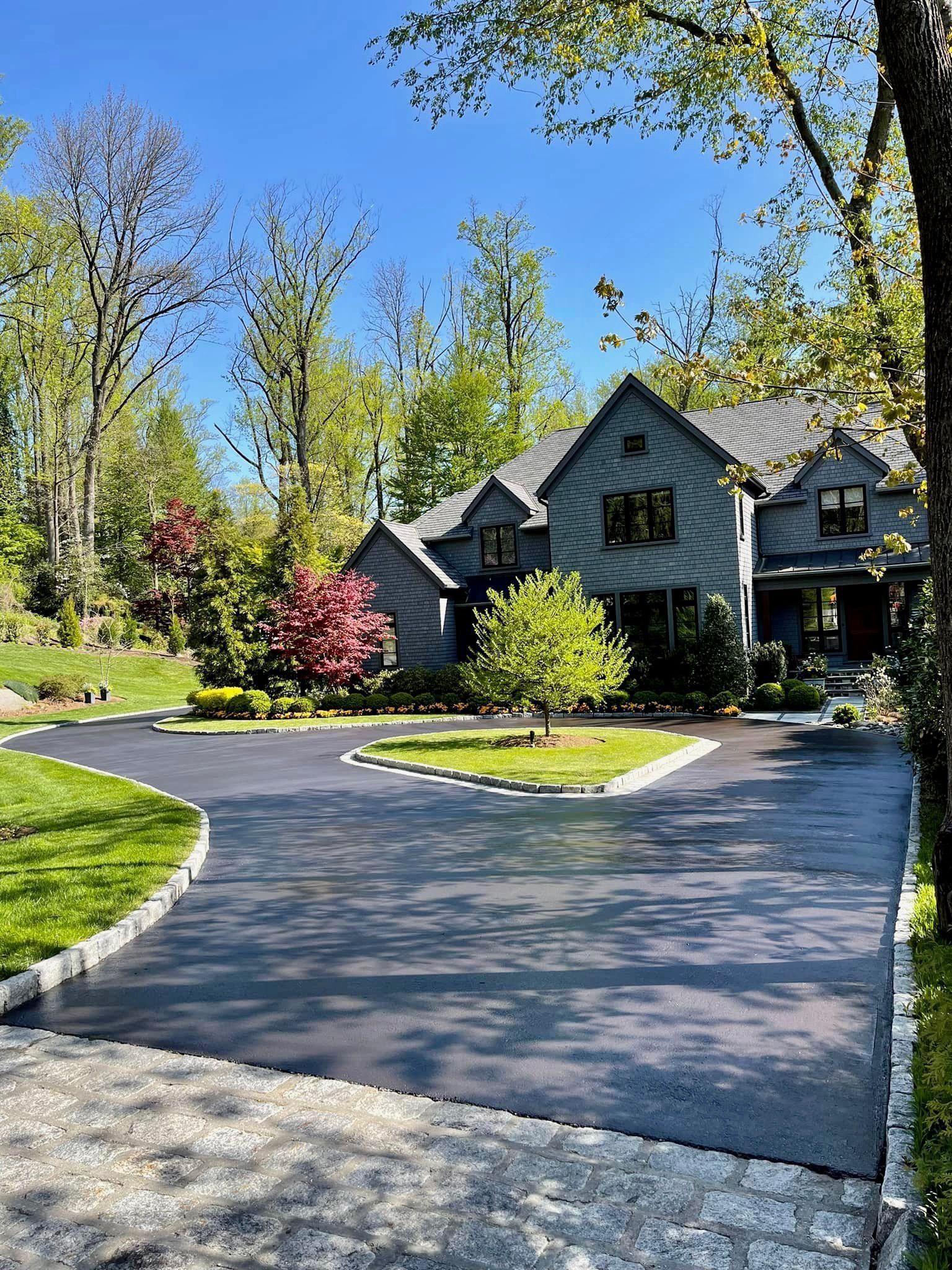 A gray suburban home with a paved driveway and a central grass island, surrounded by trees under a clear blue sky.