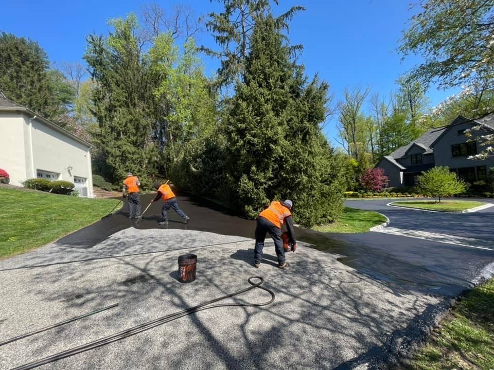 Three workers in orange safety vests apply asphalt sealer to a residential driveway on a sunny day.