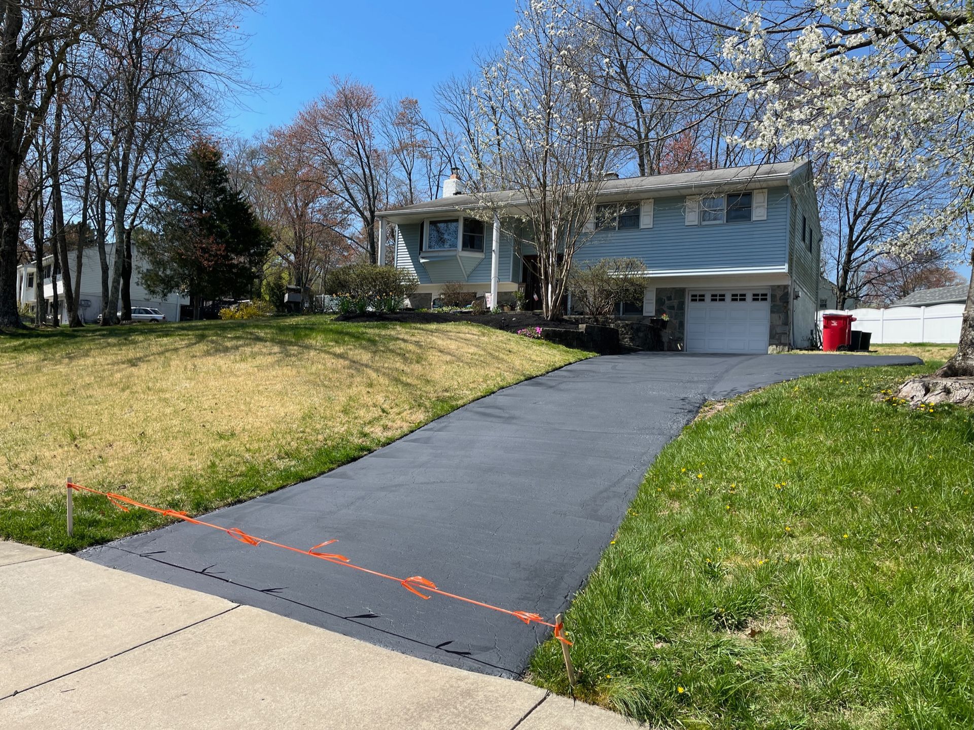 A newly paved, dark asphalt driveway leads to a light blue split-level house on a sunny day with trees in the background.