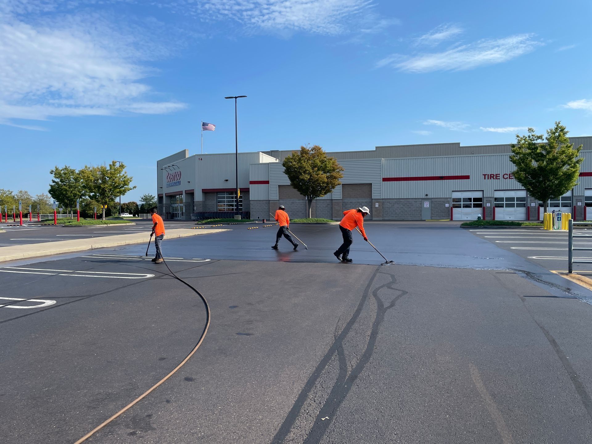 Three workers in orange high-visibility shirts sealcoating a parking lot on a sunny day.