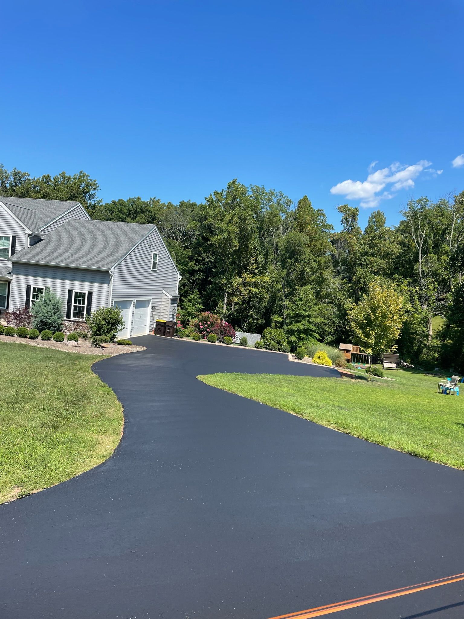 A newly paved dark asphalt driveway curves toward a suburban home surrounded by trees under a clear blue sky.