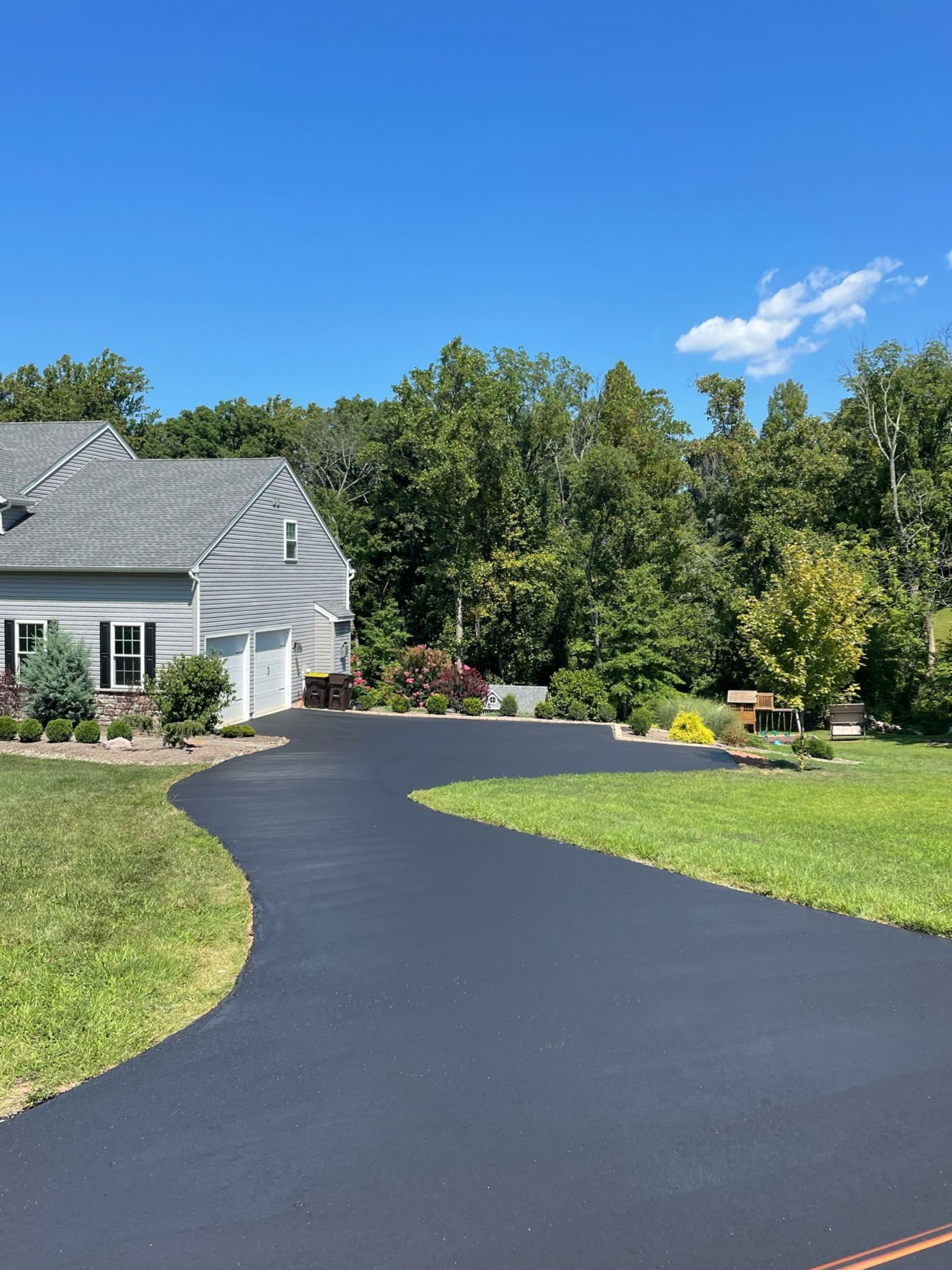 A newly paved dark asphalt driveway curves toward a suburban home surrounded by trees under a clear blue sky.