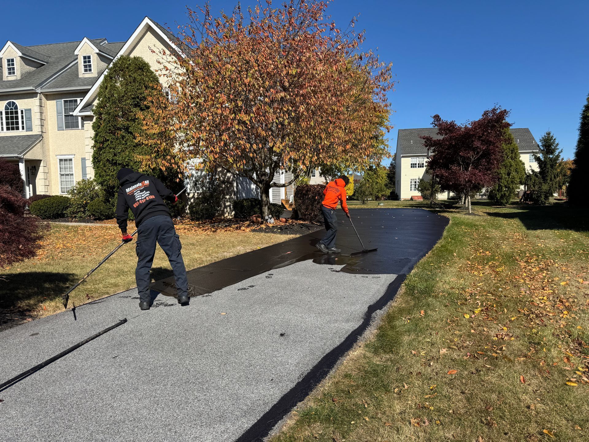 Two workers wearing high-visibility jackets apply black driveway sealant to a gravel surface on a sunny autumn day.
