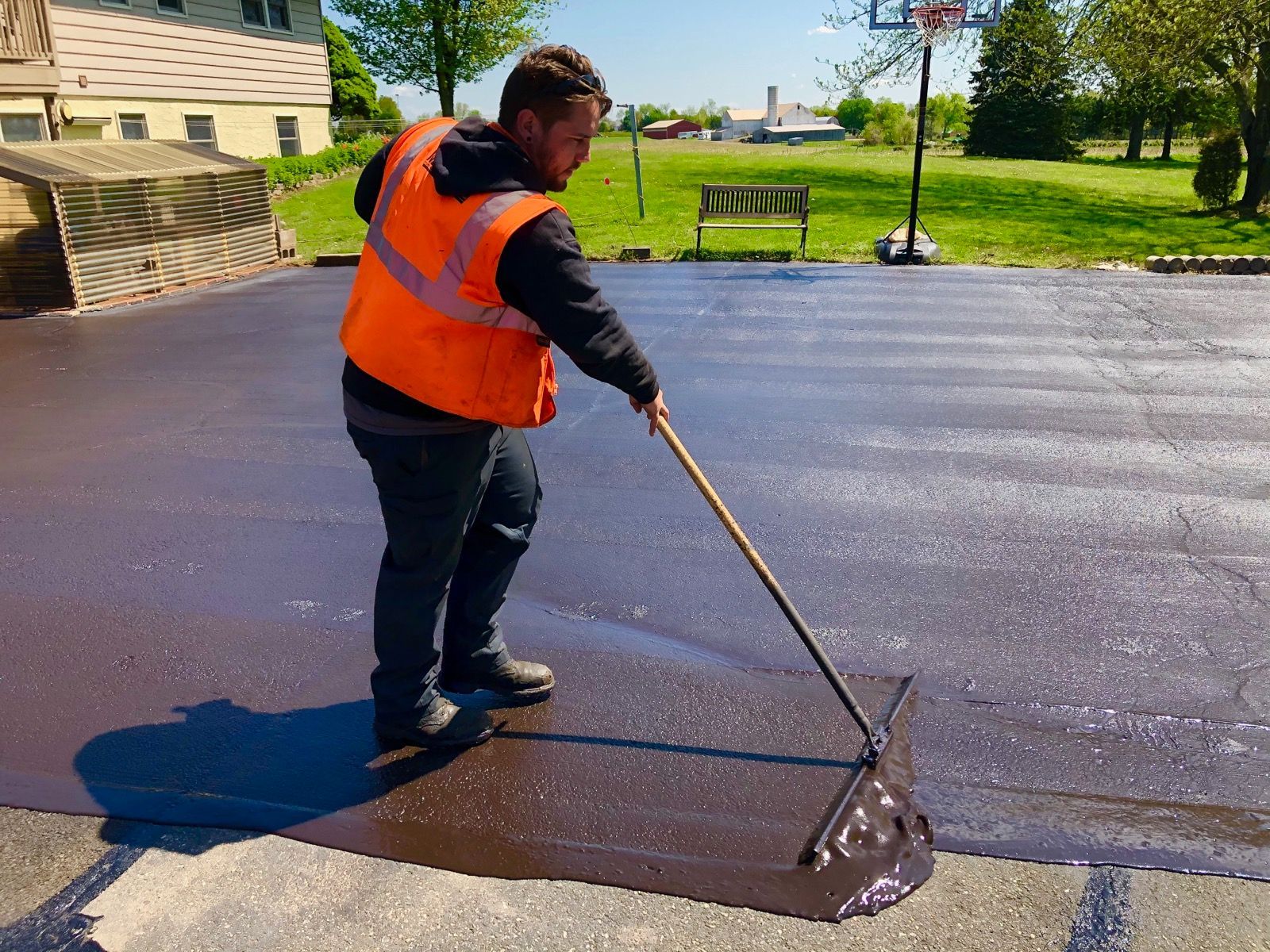 A worker wearing an orange high-visibility vest uses a squeegee to apply black asphalt sealer to a residential driveway.