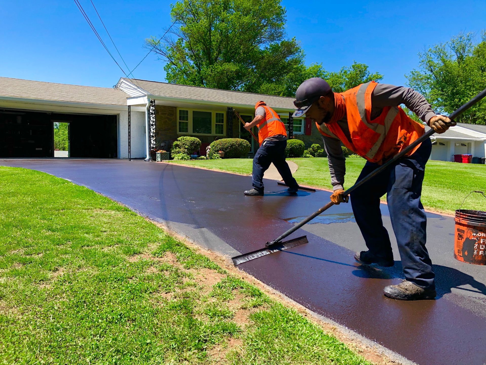 Two workers in high-visibility orange vests use squeegees to apply dark sealant to a suburban driveway on a sunny day.