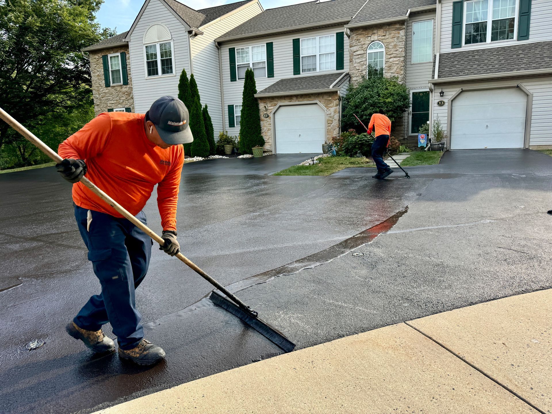 Two workers in orange shirts use squeegees to spread sealant over a residential asphalt driveway.