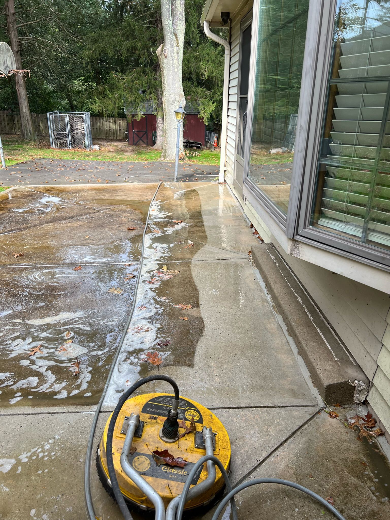 A yellow surface cleaner sits on a damp, partially power-washed concrete patio next to the exterior of a house.