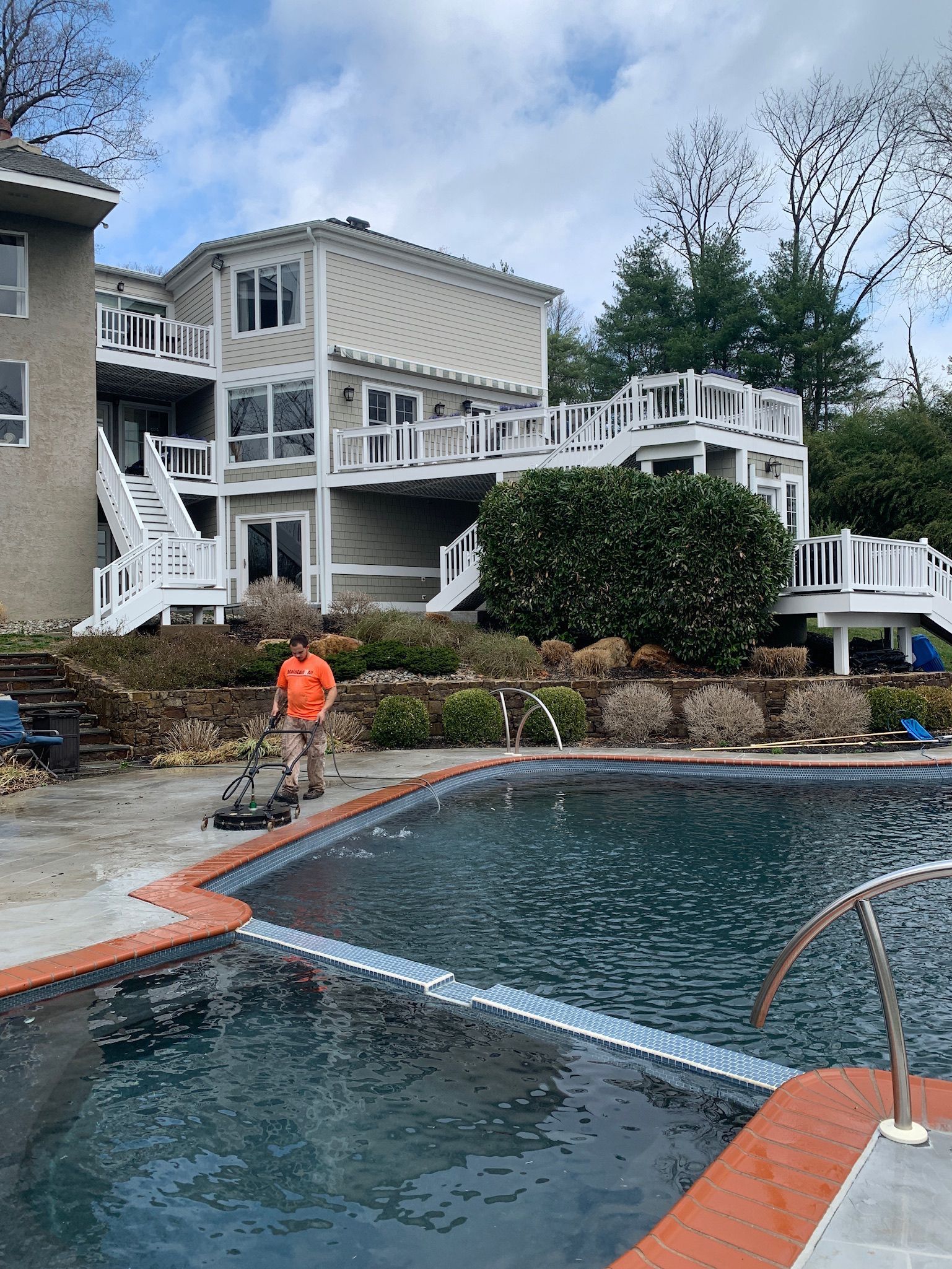 A person cleaning a patio next to a swimming pool, with a multi-level house and white railings in the background.