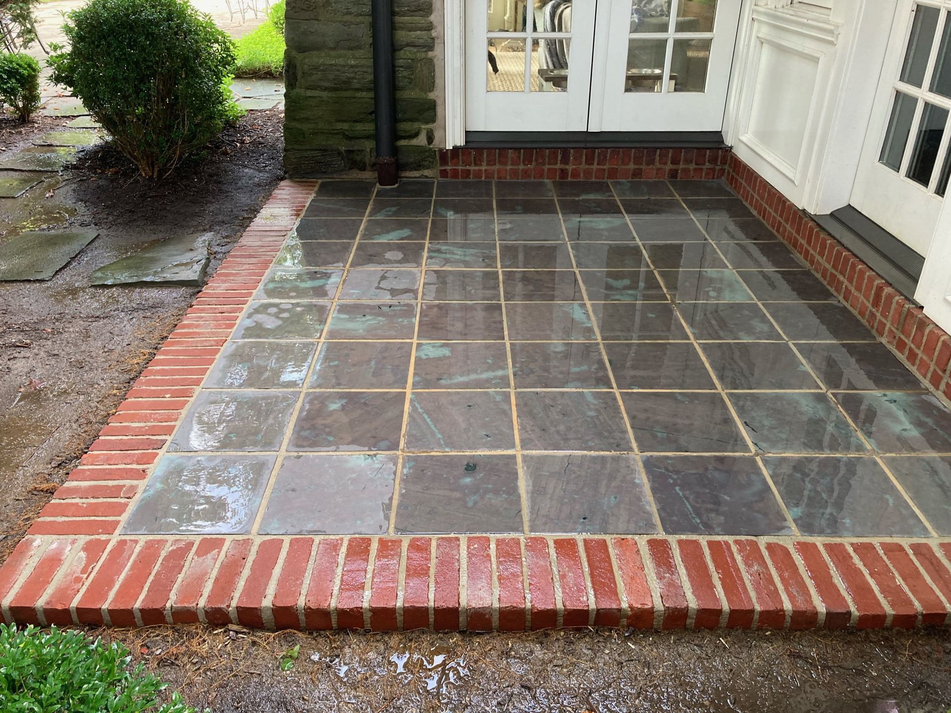 A rectangular stone patio with a brick border, adjacent to white double doors and a stone exterior wall.
