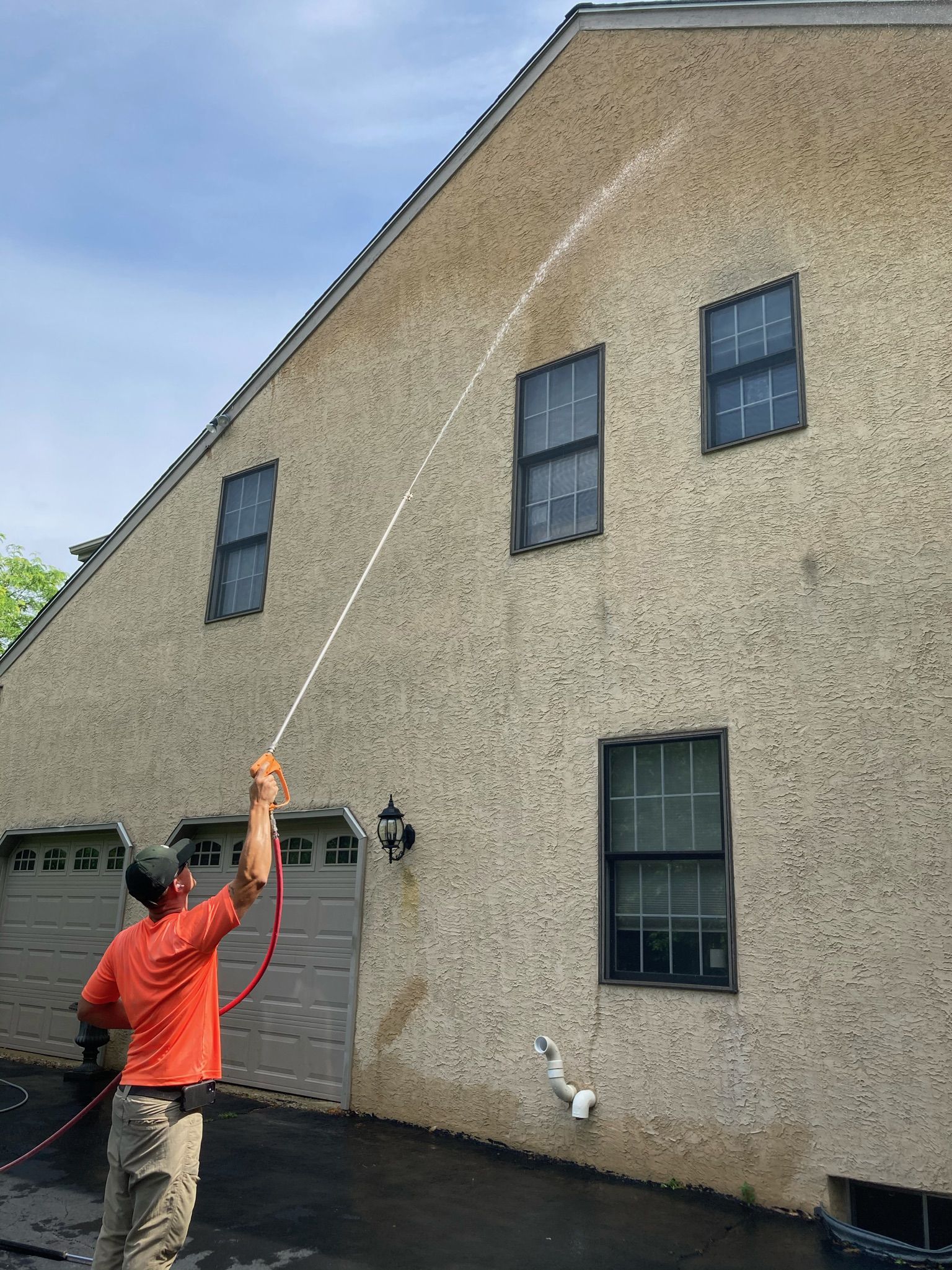 A worker in an orange shirt uses a high-pressure hose to spray clean the side of a two-story beige stucco house.