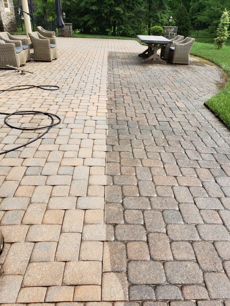 A patio undergoing power washing, showing a clear contrast between the clean, light-colored pavers and the dirty, dark ones.
