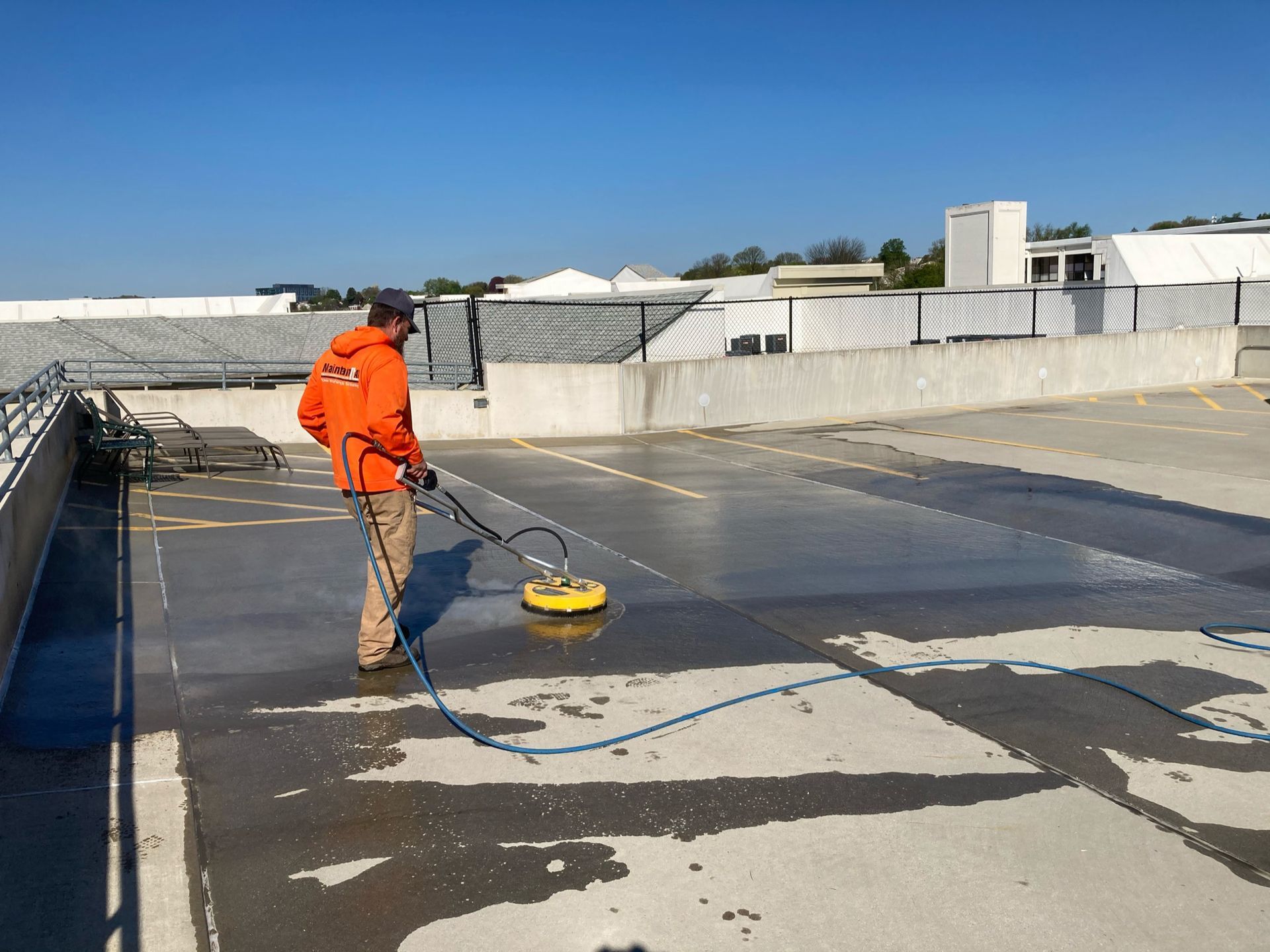 A worker in an orange jacket uses a circular pressure washer on a concrete rooftop under a clear blue sky.