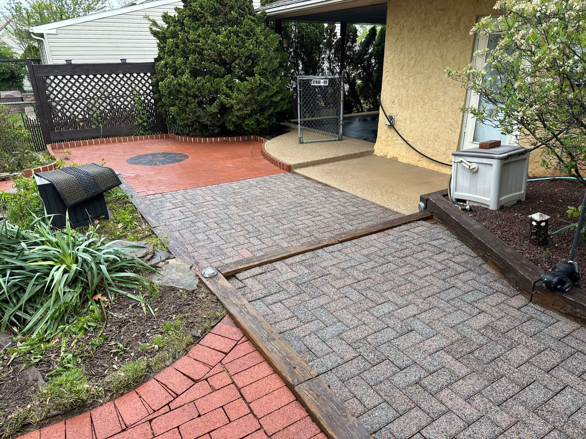 A path with red bricks and grey interlocking pavers leads to a yellow house entrance with a brown lattice fence.