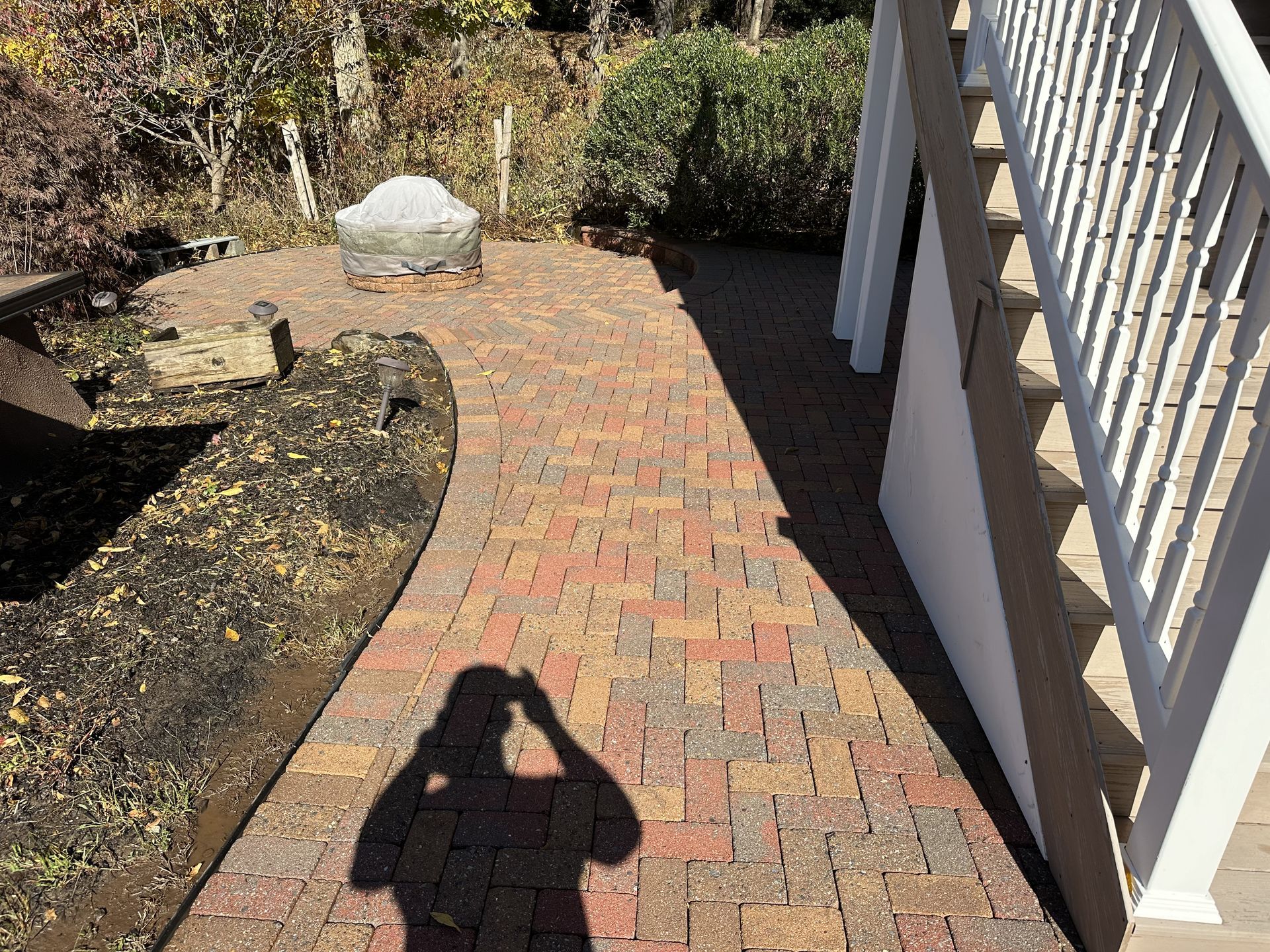 A red and tan brick walkway winds past a garden bed next to white stairs, with a photographer's shadow cast on the path.