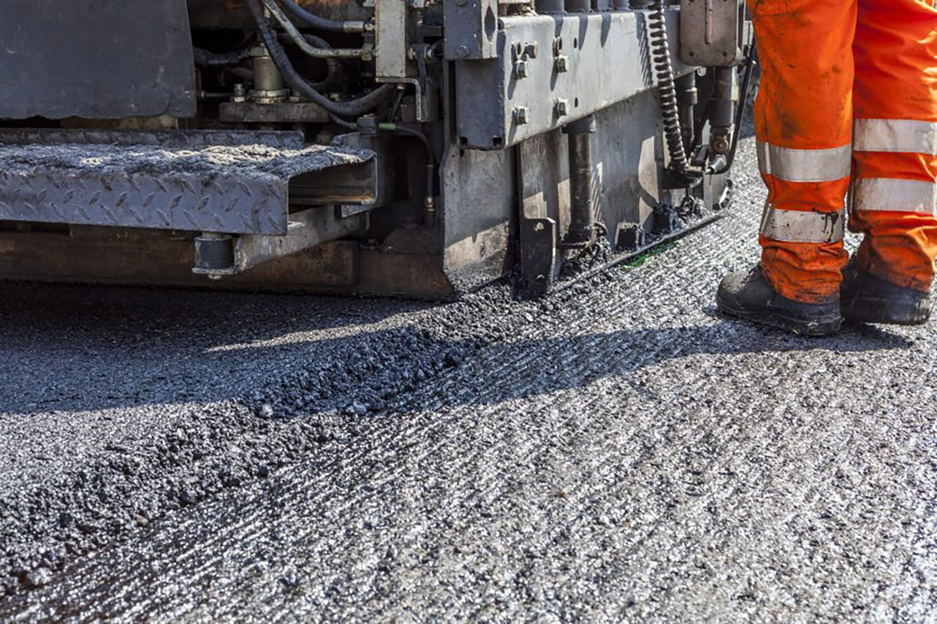 Person in orange work pants and boots near a road paving machine, laying down fresh asphalt.