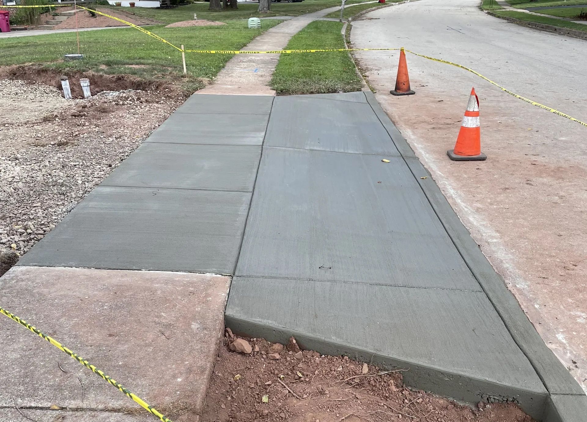 Newly poured concrete sidewalk apron with caution tape and orange cones in a residential area.