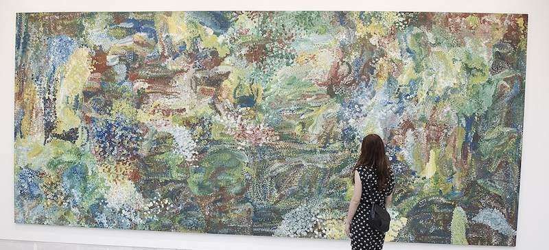 A person stands before a large, horizontal abstract painting filled with colorful, textured, and expressive brushstrokes.