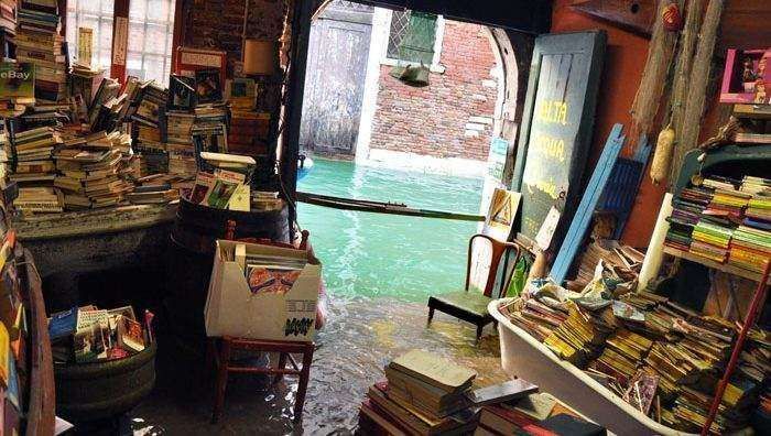 Interior of a bookstore in Venice, Italy, with shelves of books and water flooding the floor, leading to a canal outside.