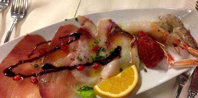 A plate of assorted raw seafood carpaccio garnished with pomegranate seeds, balsamic glaze, and an orange slice.