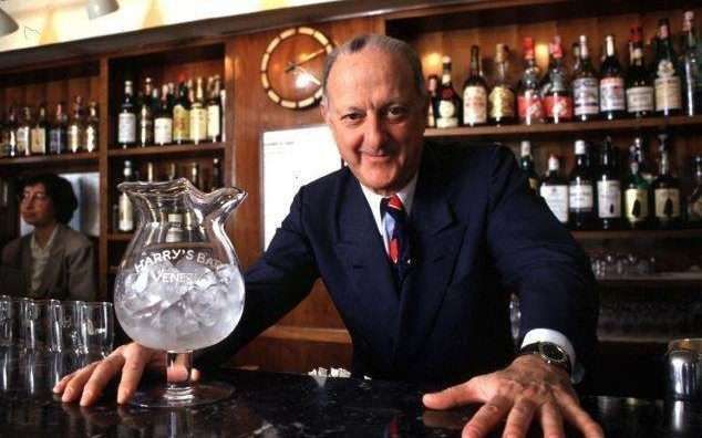 A smiling person in a suit stands behind a bar with a pitcher of ice, set against shelves filled with liquor bottles.